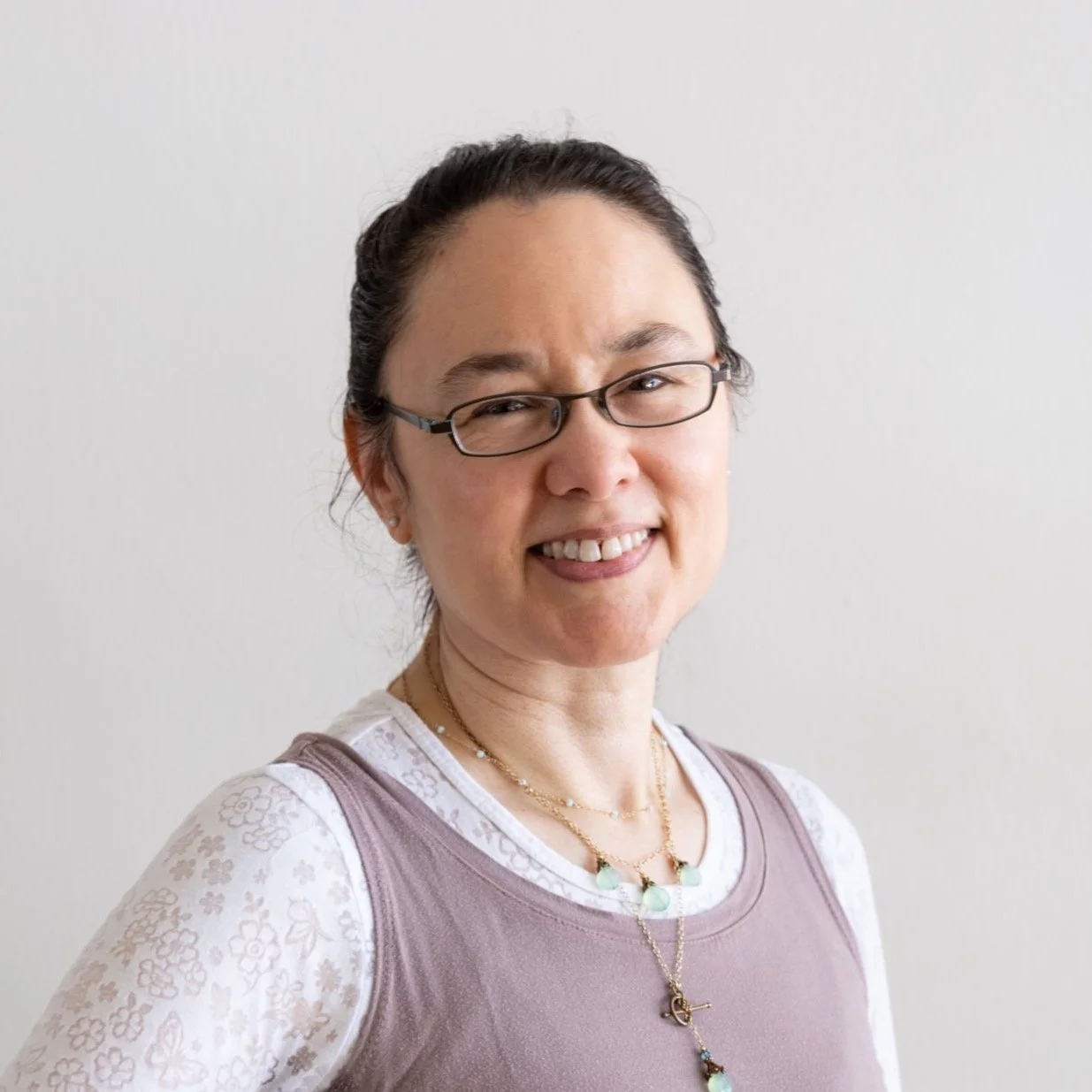 Portrait of a woman with glasses, smiling, wearing a floral-patterned white shirt and layered necklaces, against a plain background.
