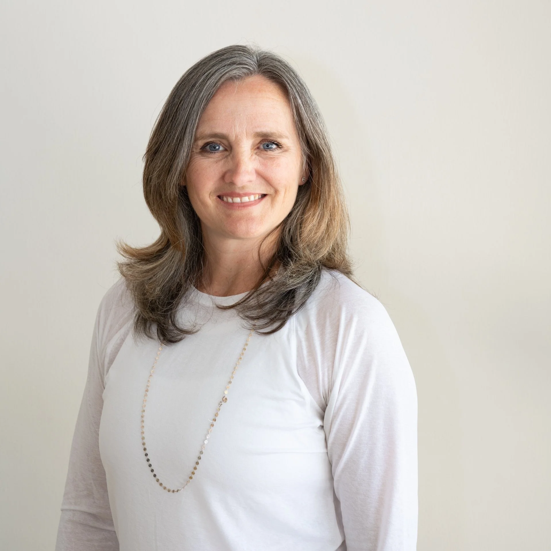 A middle-aged woman with shoulder-length gray hair, blue eyes, and a warm smile, wearing a white top and a delicate beaded necklace, standing against a plain light background.