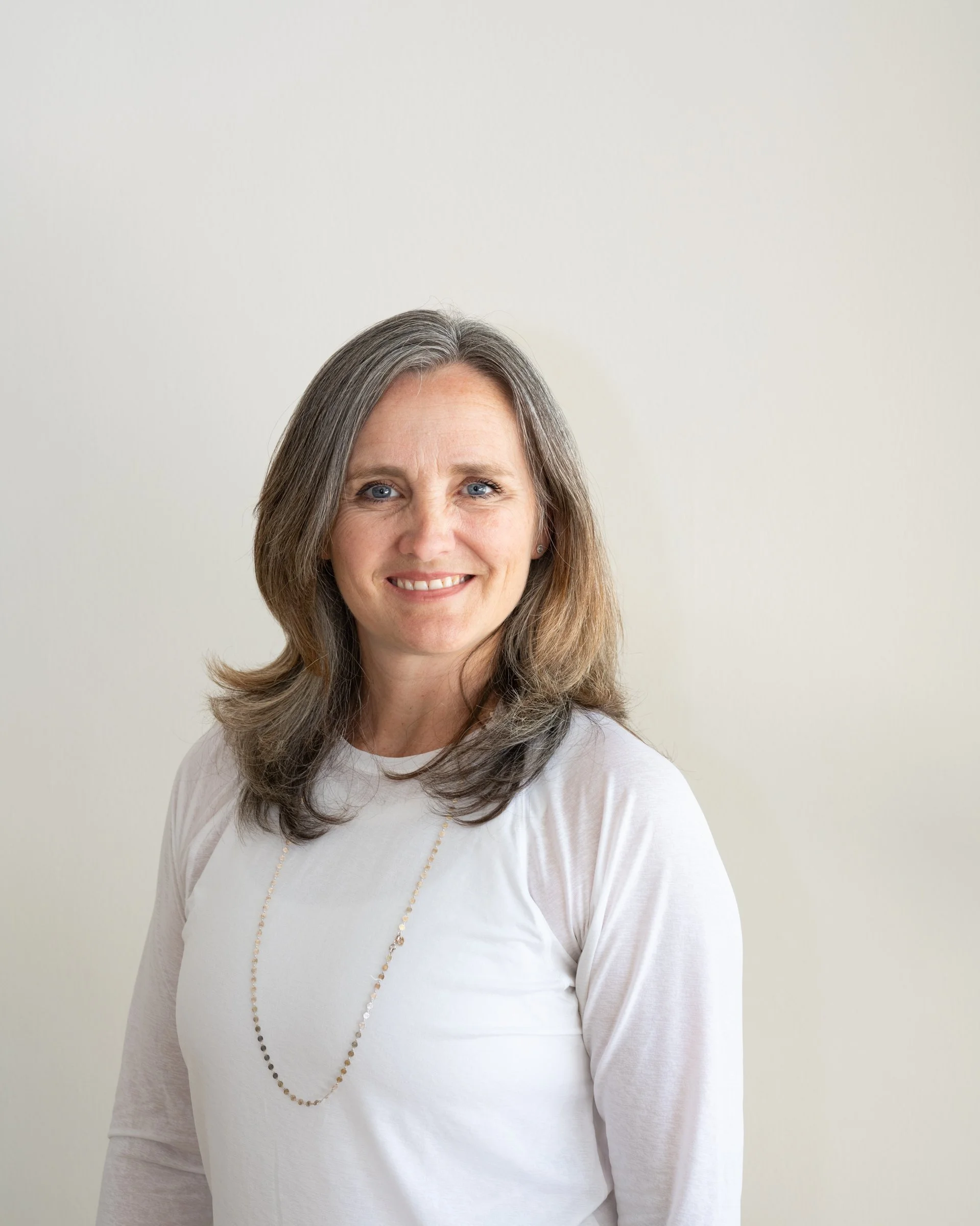 A middle-aged woman with shoulder-length brown hair, blue eyes, and a warm smile, wearing a white long-sleeve top and a beaded necklace, standing against a plain off-white background.
