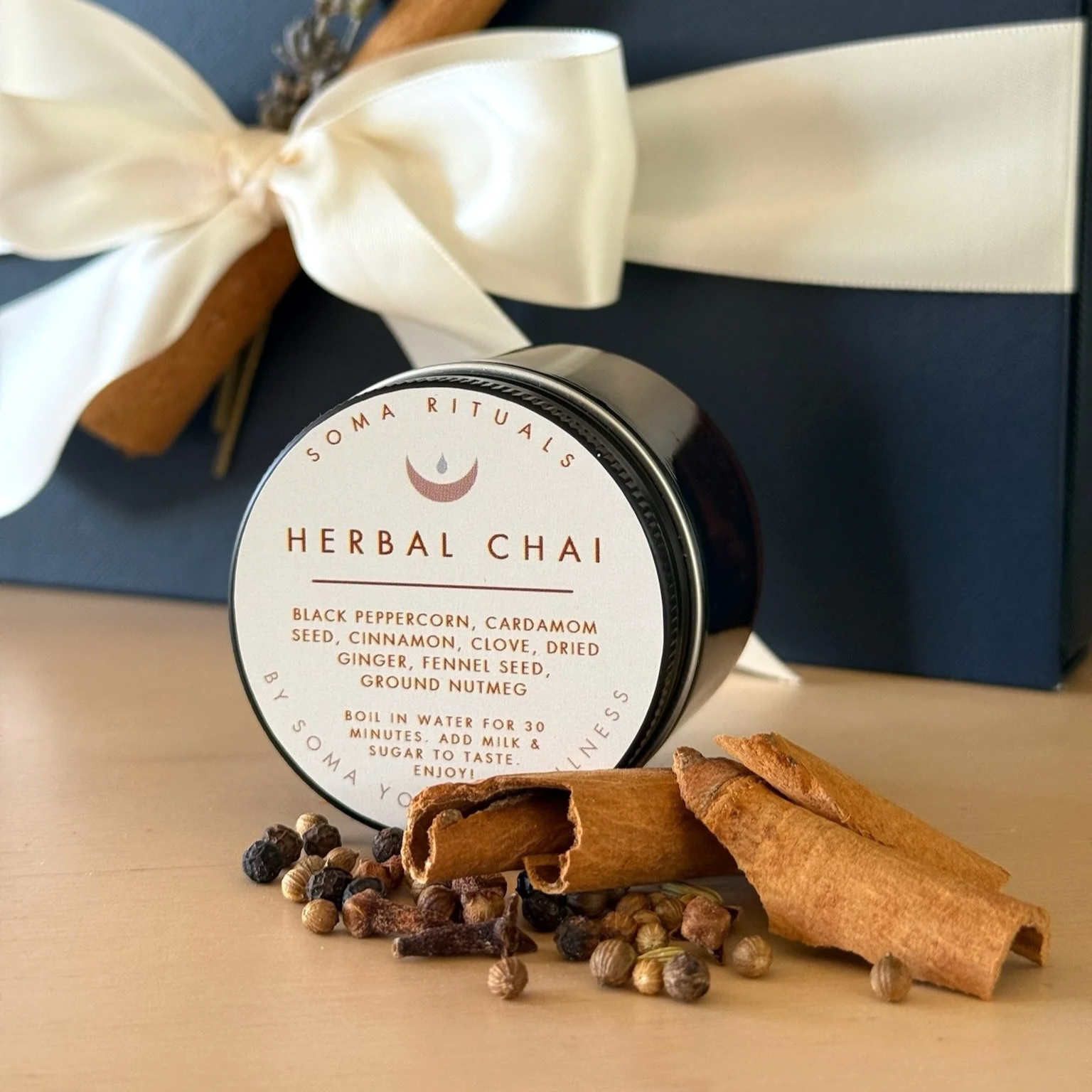 A tin container labeled 'Herbal Chai' from Soma Rituals, along with cinnamon sticks, black peppercorns, and coriander seeds on a light-colored surface, with a dark blue gift box and a cream-colored ribbon in the background.