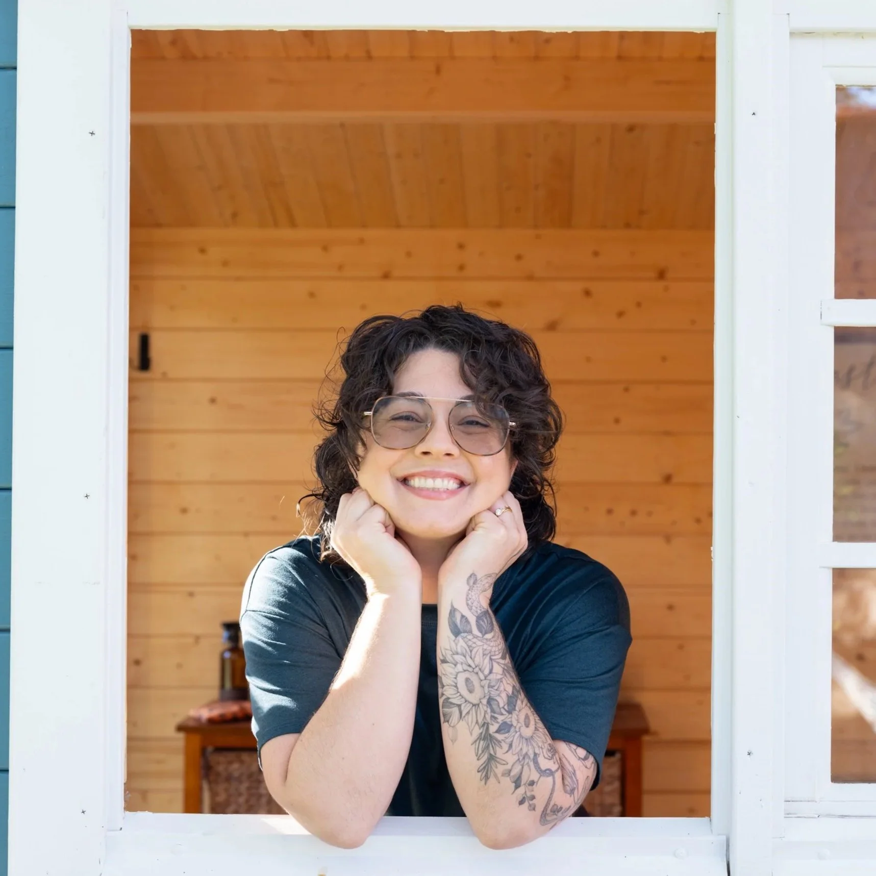 A woman with short, curly hair, glasses, and hoop earrings, smiling and wearing a light-colored shirt with dark drawstring pants, standing against a plain background.