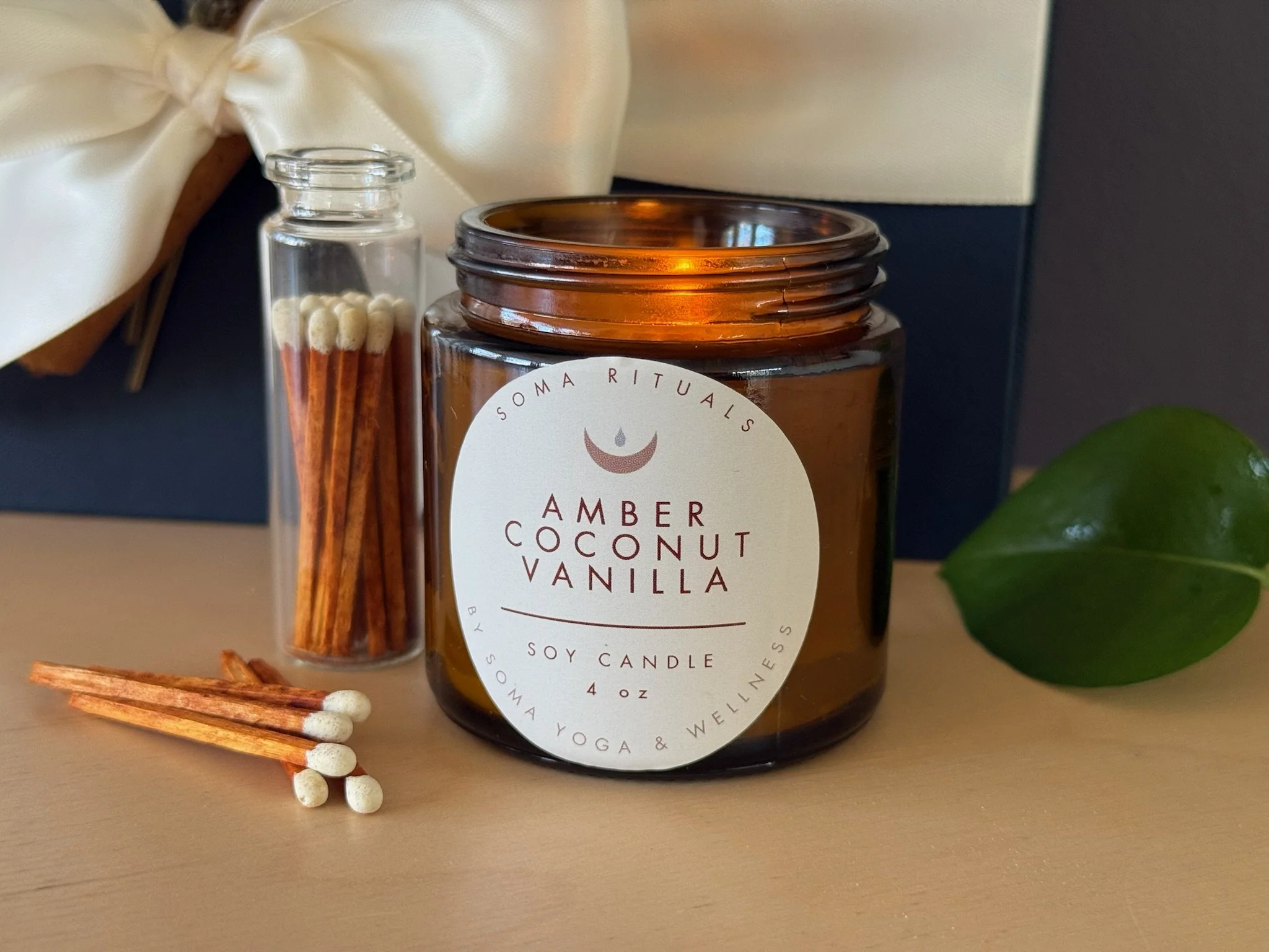 A soy candle in a brown glass jar labeled "Amber Coconut Vanilla" next to a small plastic container with cinnamon sticks and matches, a green leaf, and some matches with white tips.