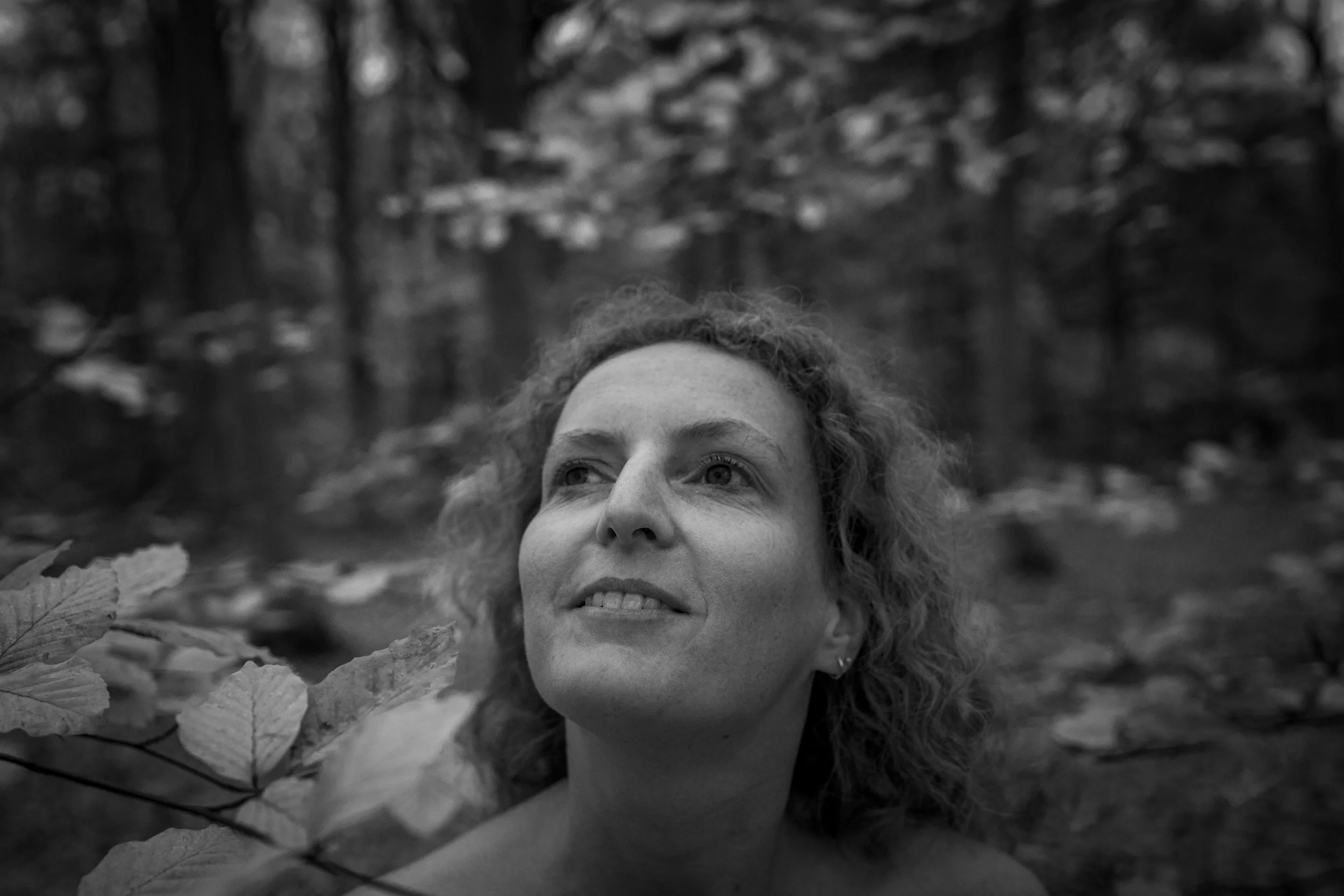Portret van een vrouw met schouderlang, krullend haar, in een bos met bladeren op de achtergrond in zwart-wit.