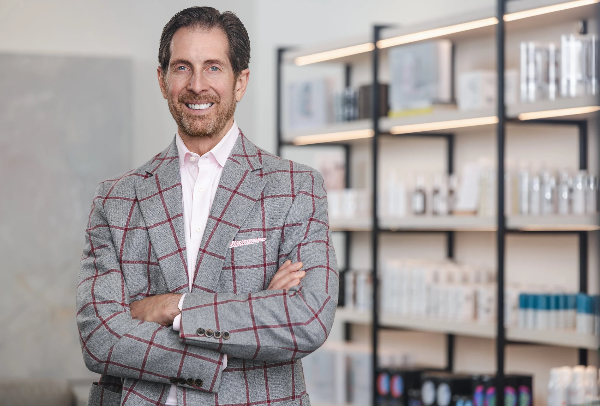 Man in a gray and red windowpane blazer standing with arms crossed in front of shelves of skincare products.