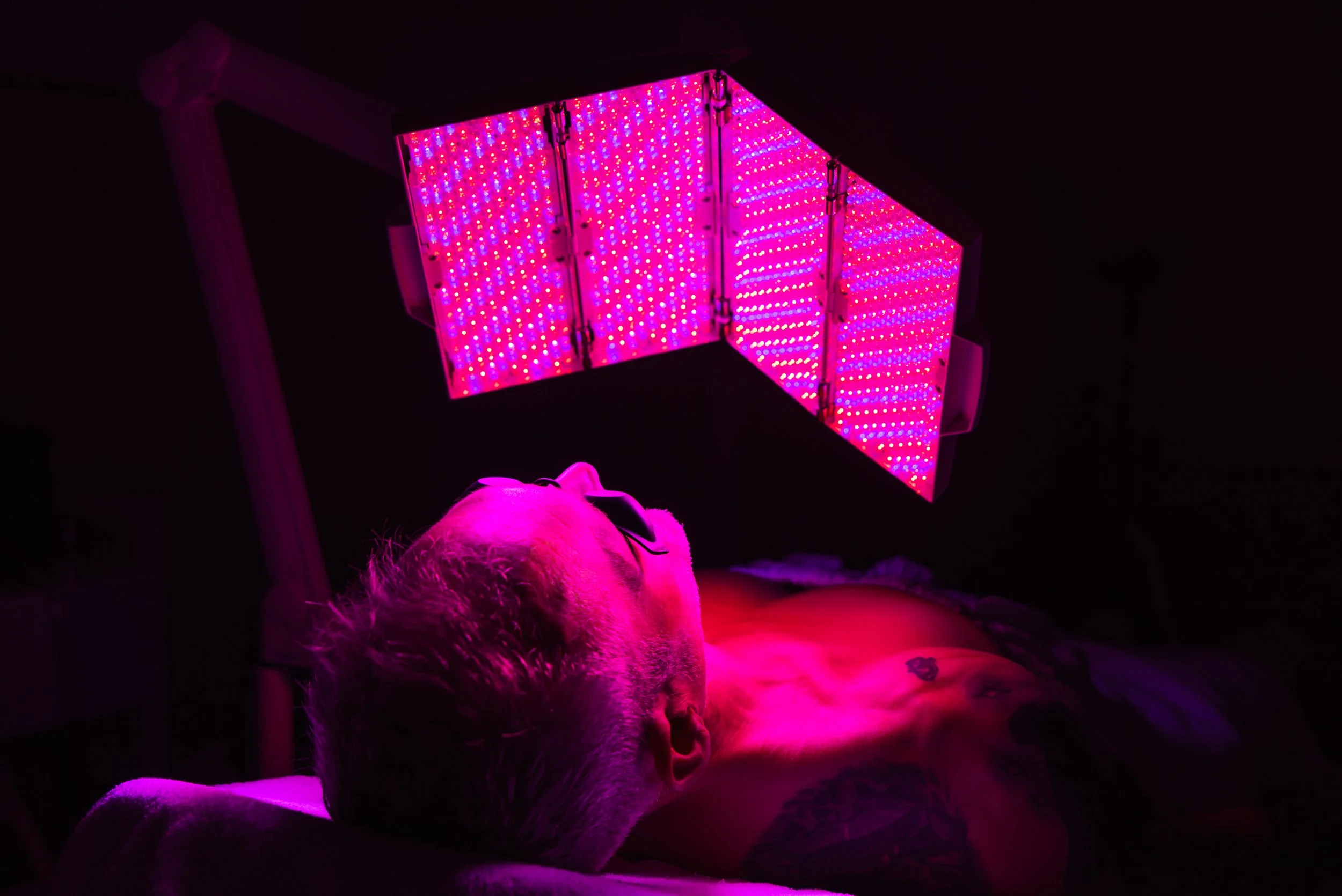 Man lying back under a red LED light therapy panel wearing protective eyewear during a skin treatment session.