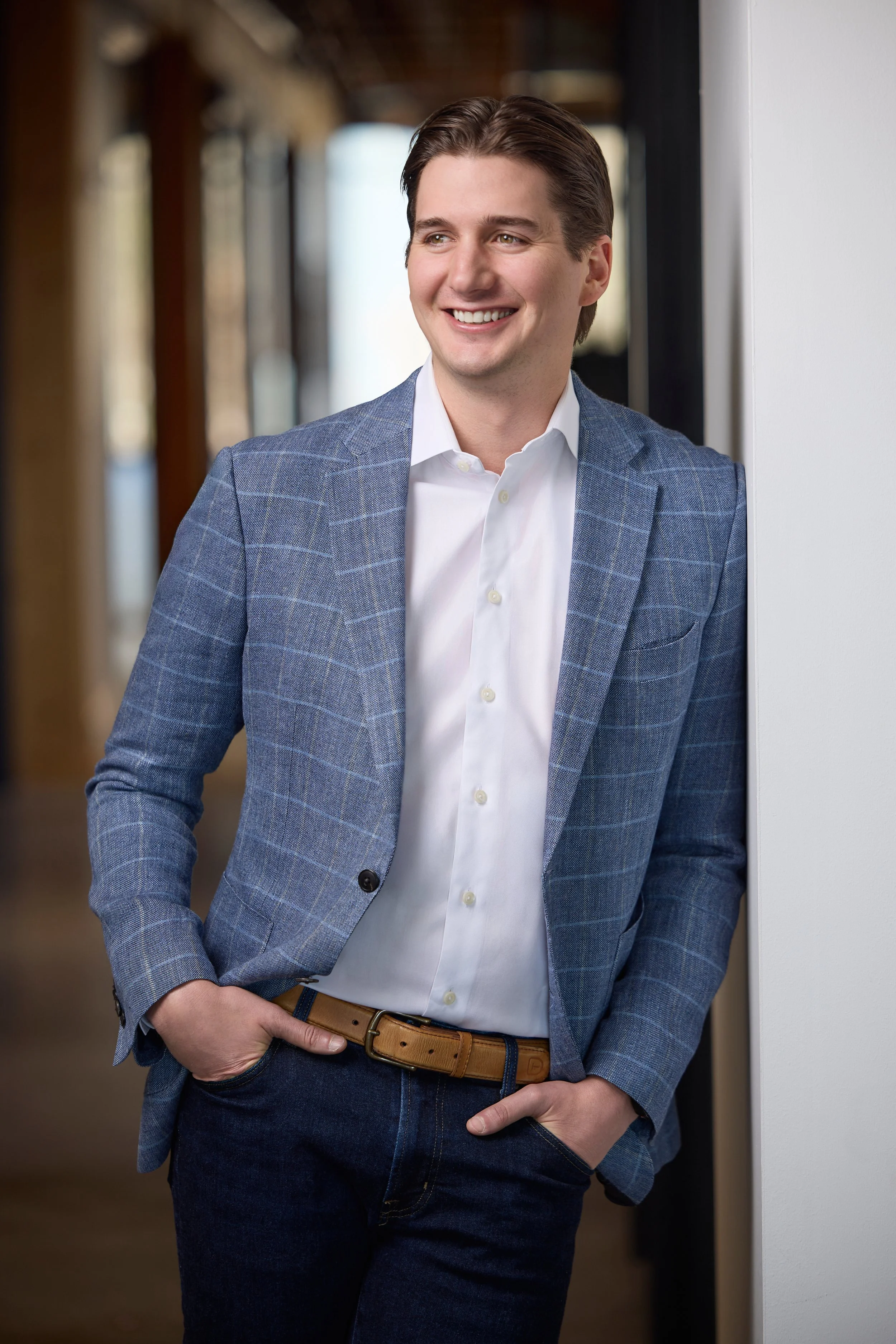 Professional headshot of a smiling man in a blue plaid blazer leaning against a wall in a warm-toned office hallway, photographed by Tausha Dickinson Photography