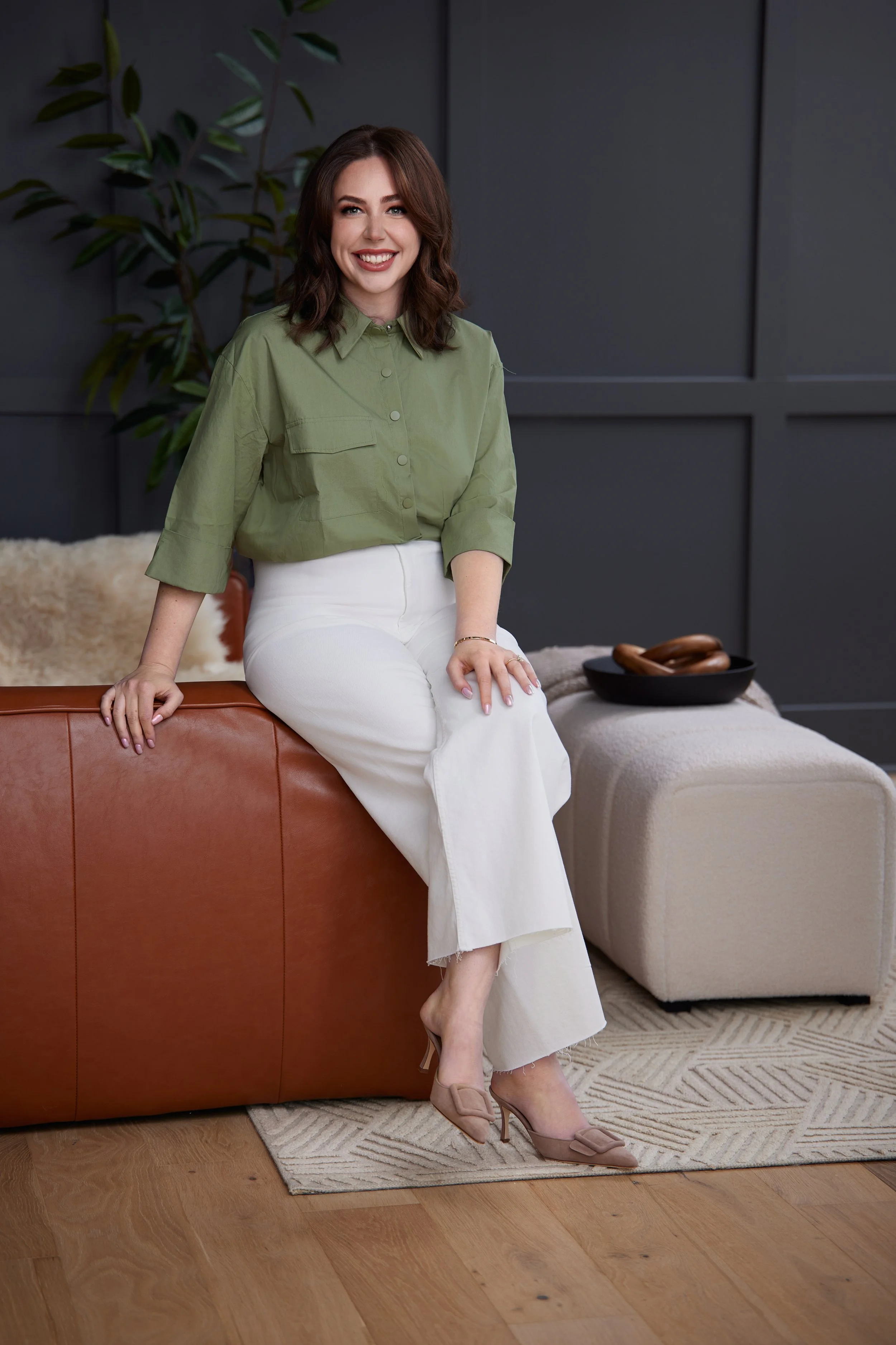 Personal branding photo of a smiling woman in a green shirt seated on a leather ottoman in a styled interior setting, photographed by Tausha Dickinson in Nashville