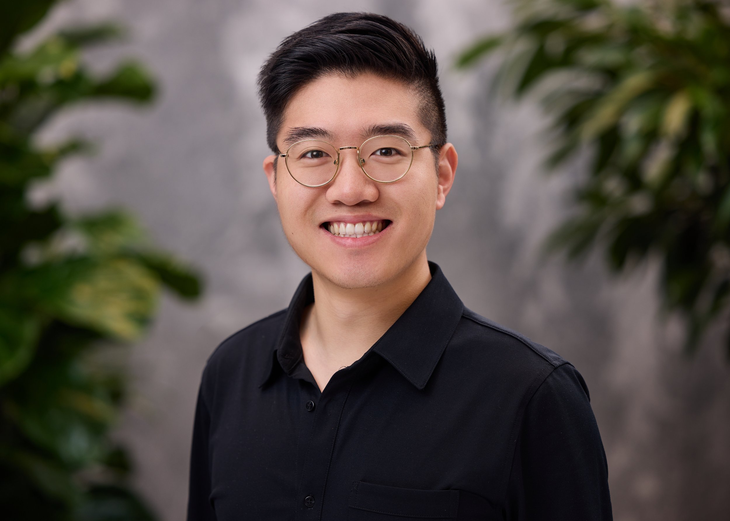 LinkedIn headshot of a smiling man with glasses in a black button-up shirt against a soft green foliage background.