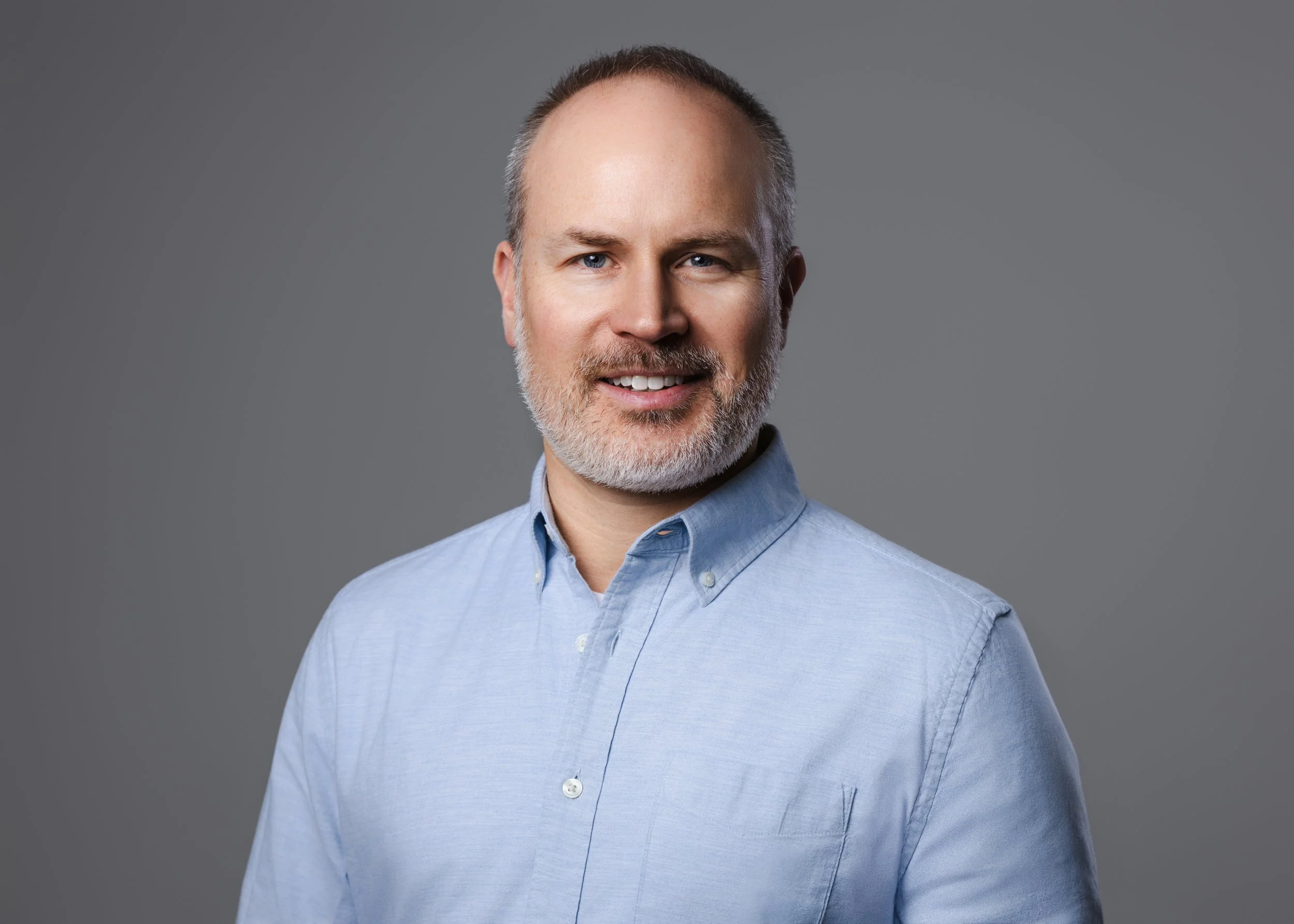 Casual professional headshot of a man with gray hair and a beard in a light blue button-down shirt.