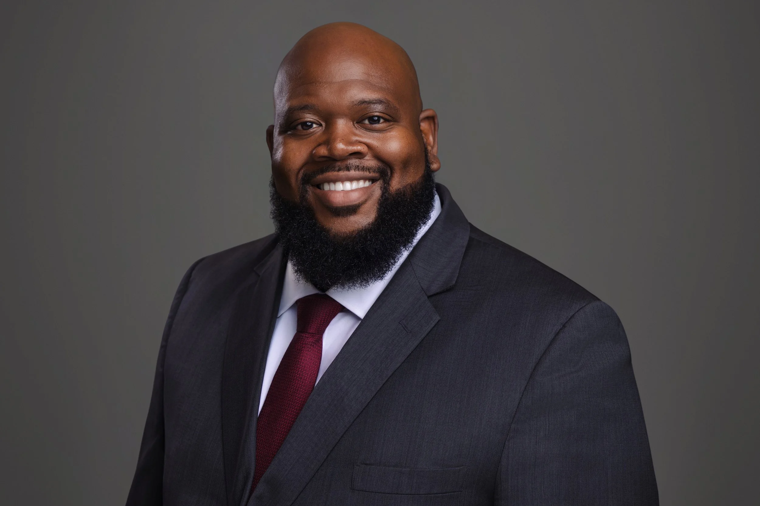 Professional headshot of a smiling man with a full beard wearing a dark suit and red tie.