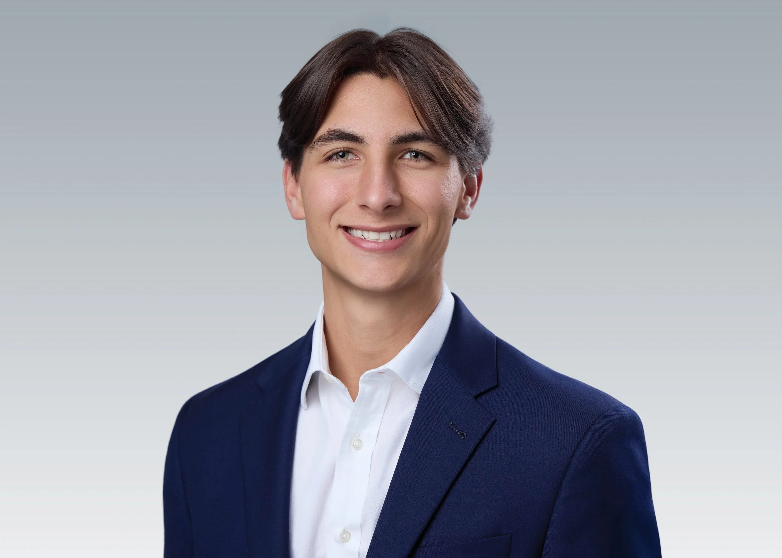 Studio headshot of a young man with clear skin in a navy blazer and white shirt.