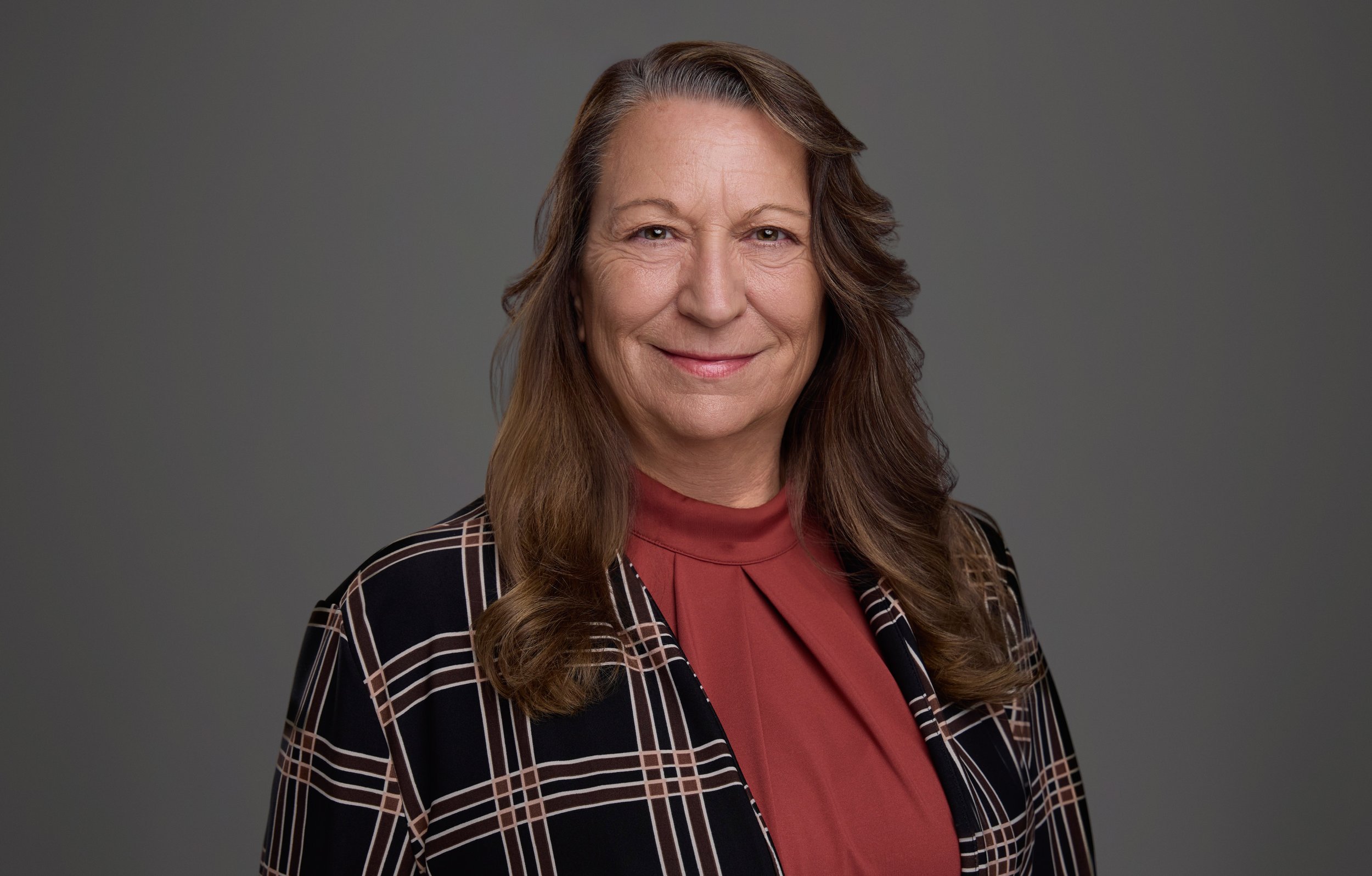 Corporate headshot of a Thompson Burton team member in a plaid jacket and rust blouse on a consistent gray studio background, by Nashville headshot photographer Tausha Dickinson