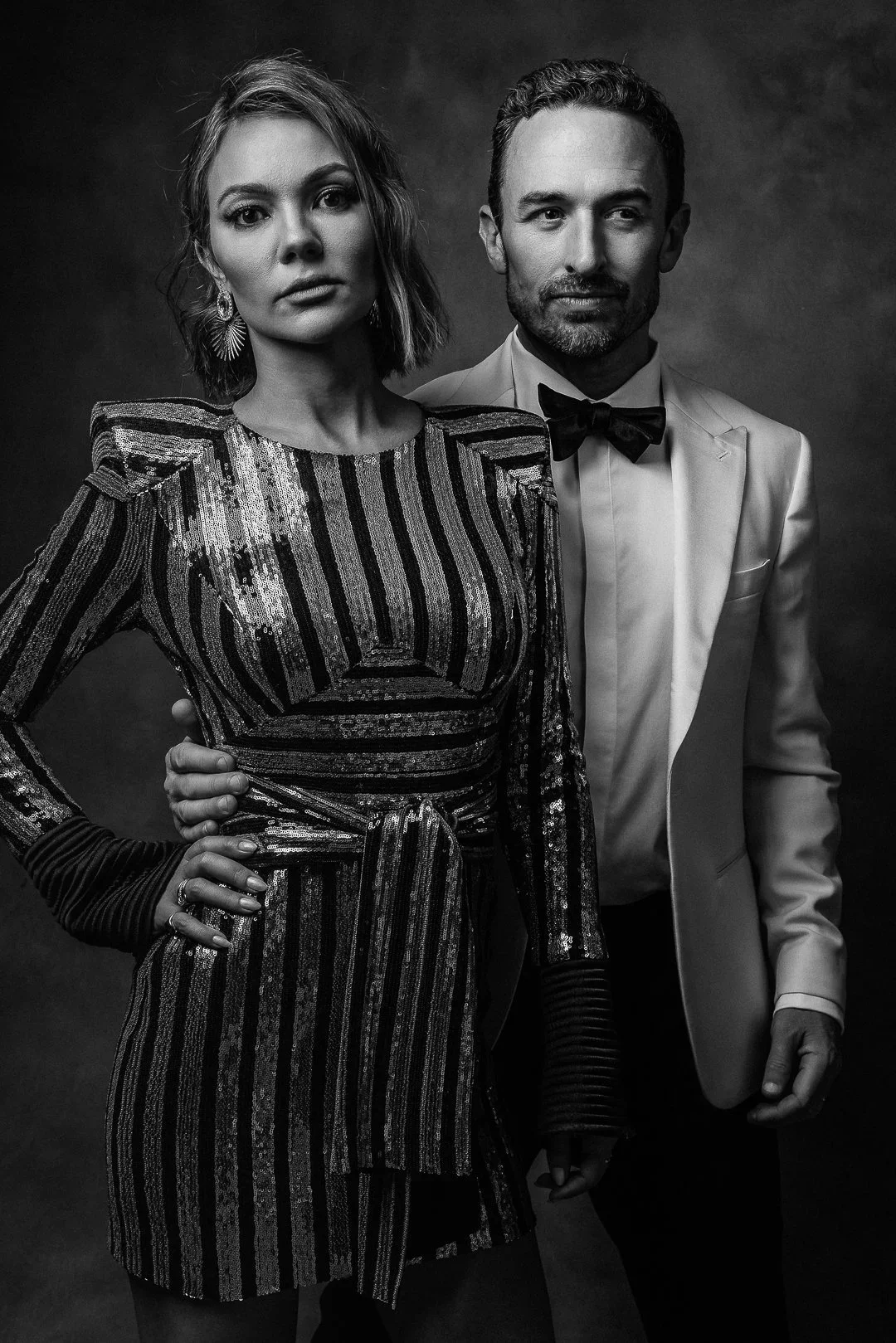 Elegant black and white event portrait of couple in formal attire - woman in sequined striped dress and man in white tuxedo with bow tie.