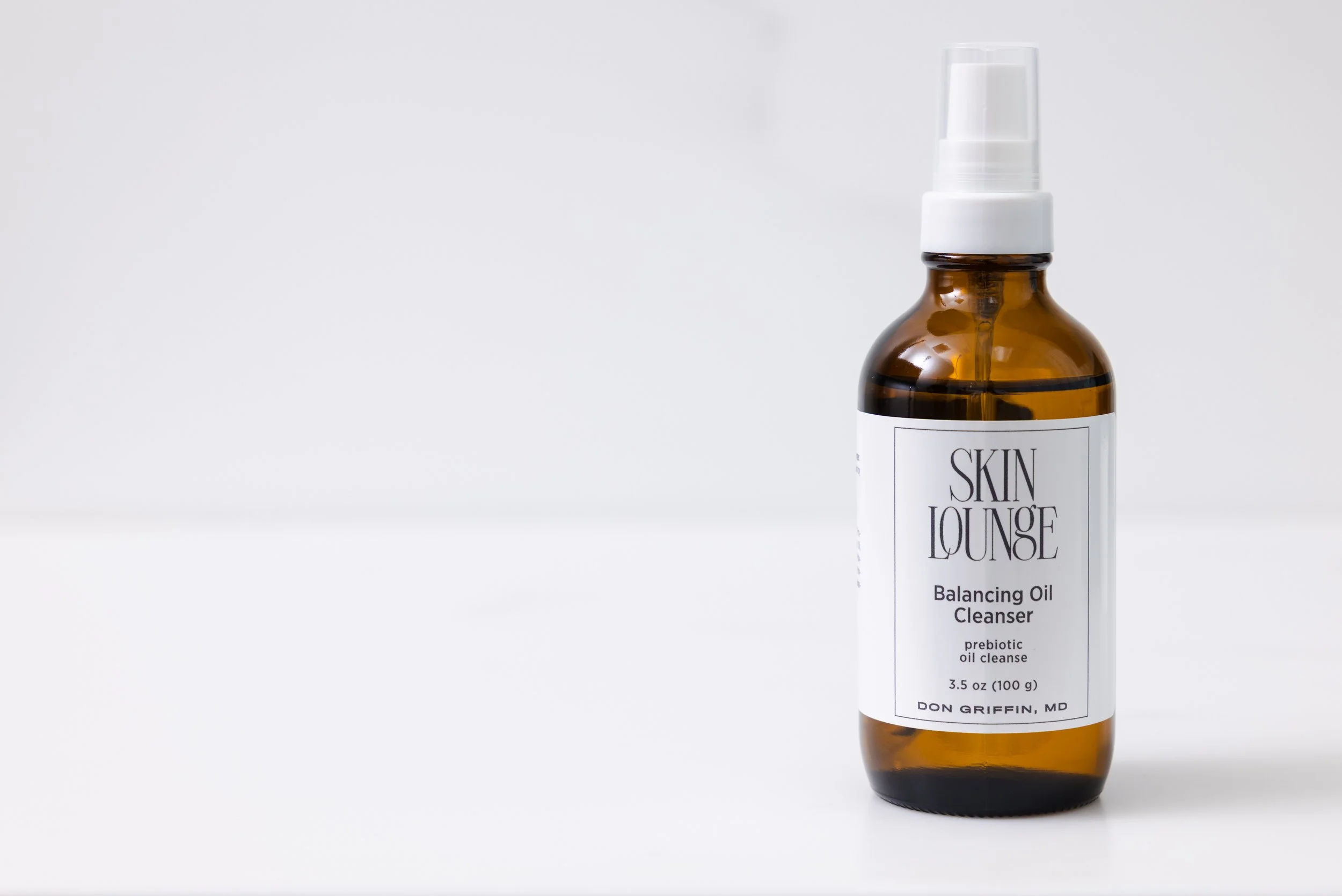 Amber glass bottle of Skin Lounge Balancing Oil Cleanser with a white pump top on a white background.