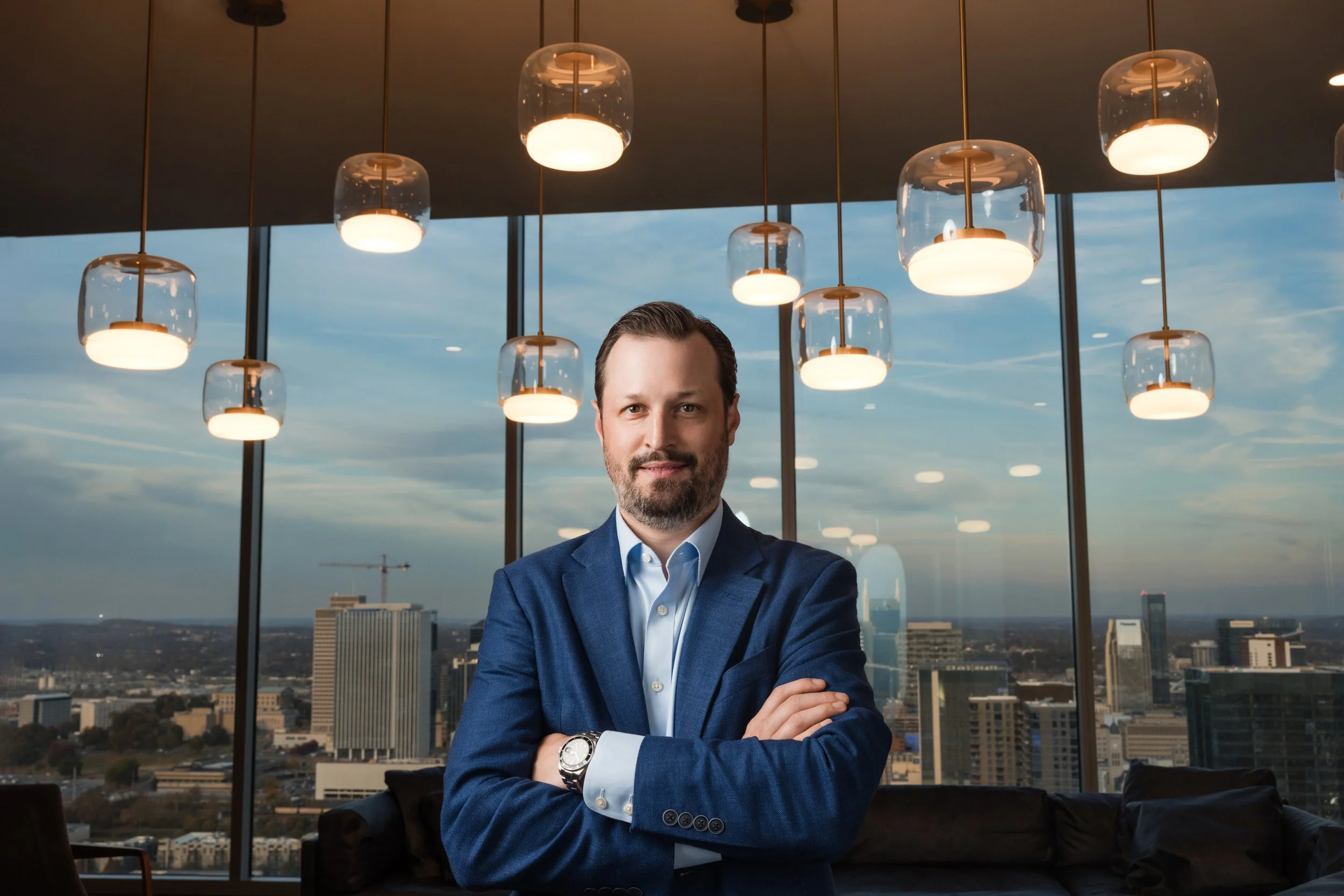 Executive headshot of a man in a blue blazer with arms crossed in front of windows overlooking the Nashville skyline.