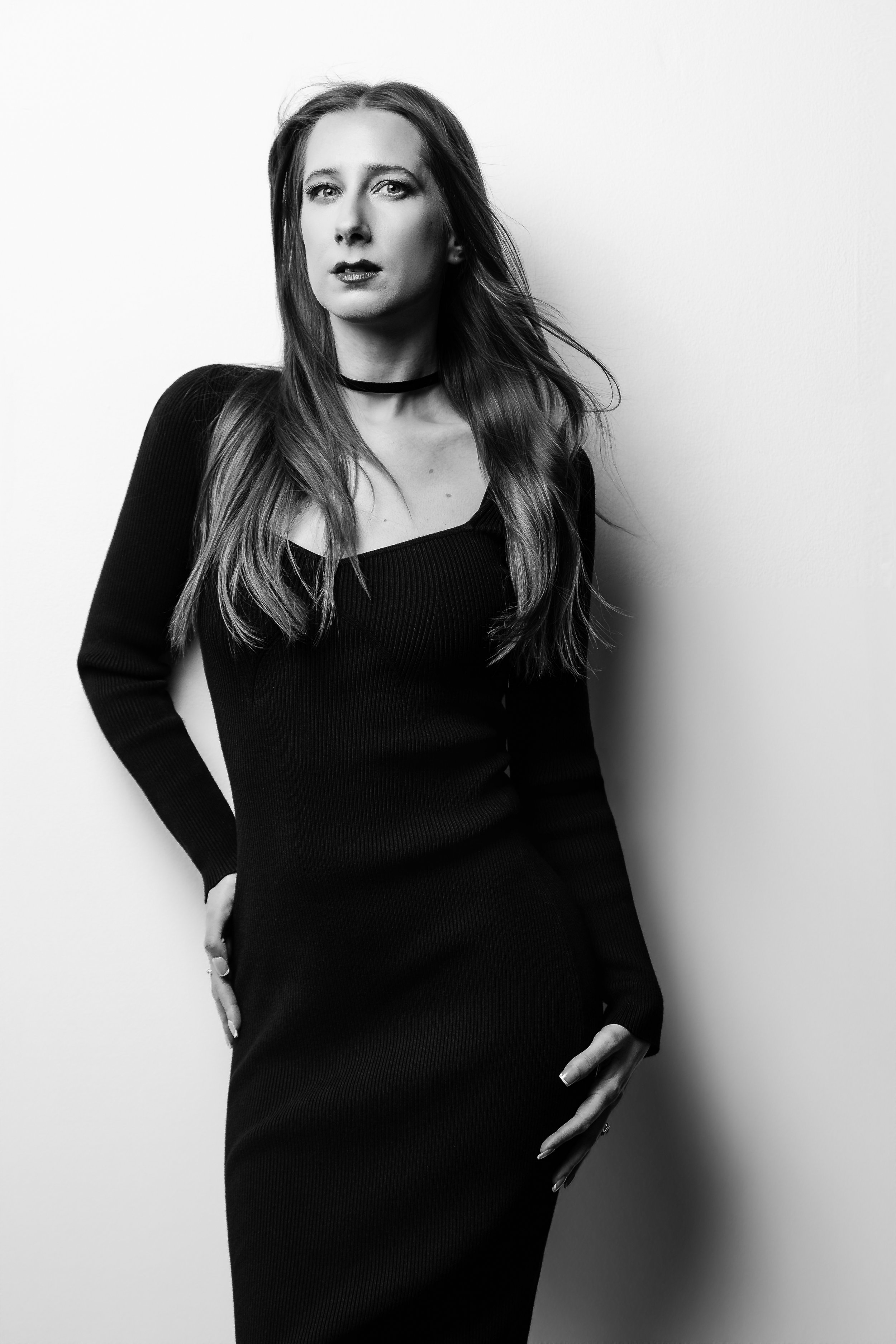 Black and white studio portrait of Lauren River leaning against a white wall with soft shadow falloff and minimal one-light portrait lighting.