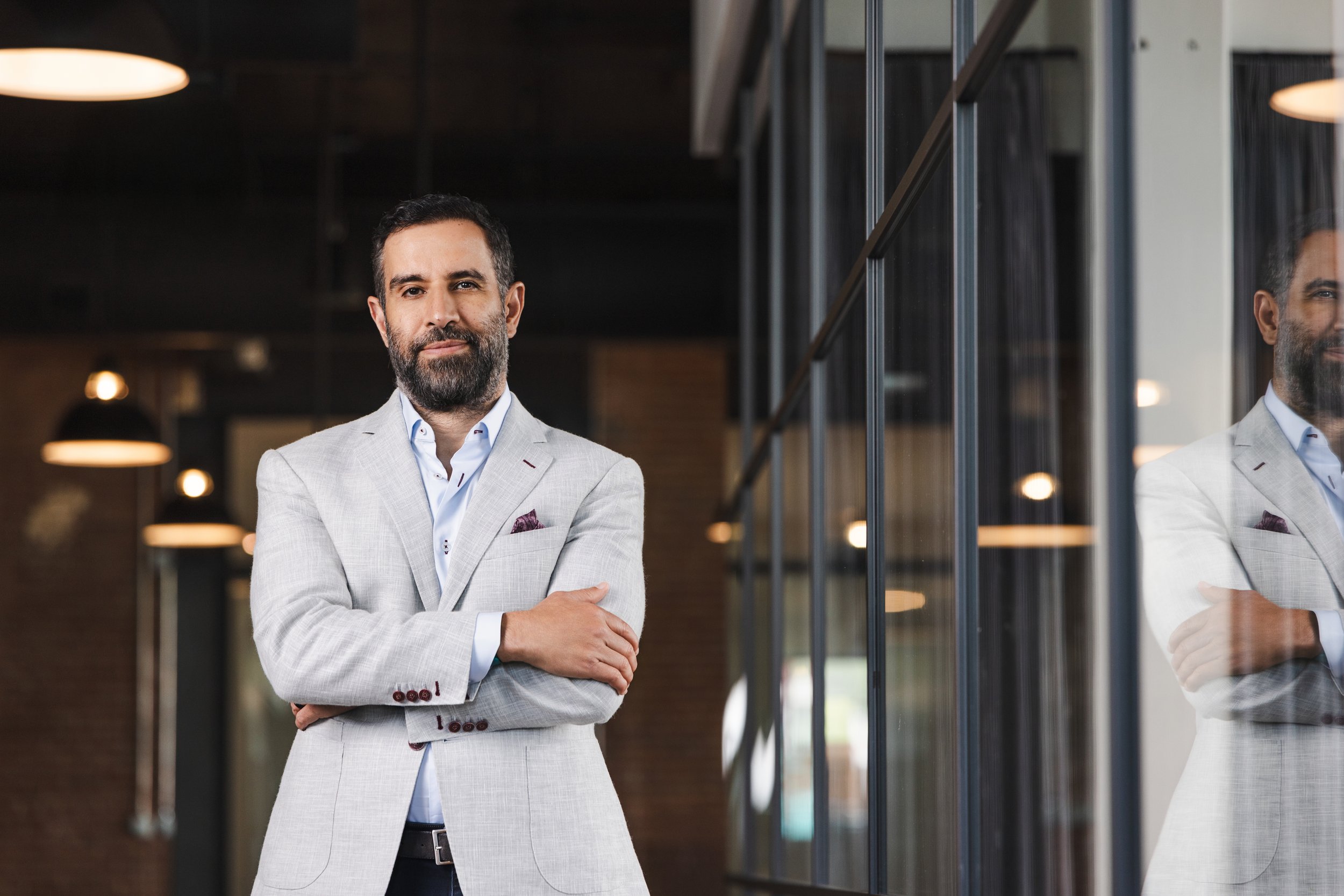 Environmental headshot of a man in a light gray blazer standing in a modern glass office with his arms crossed.