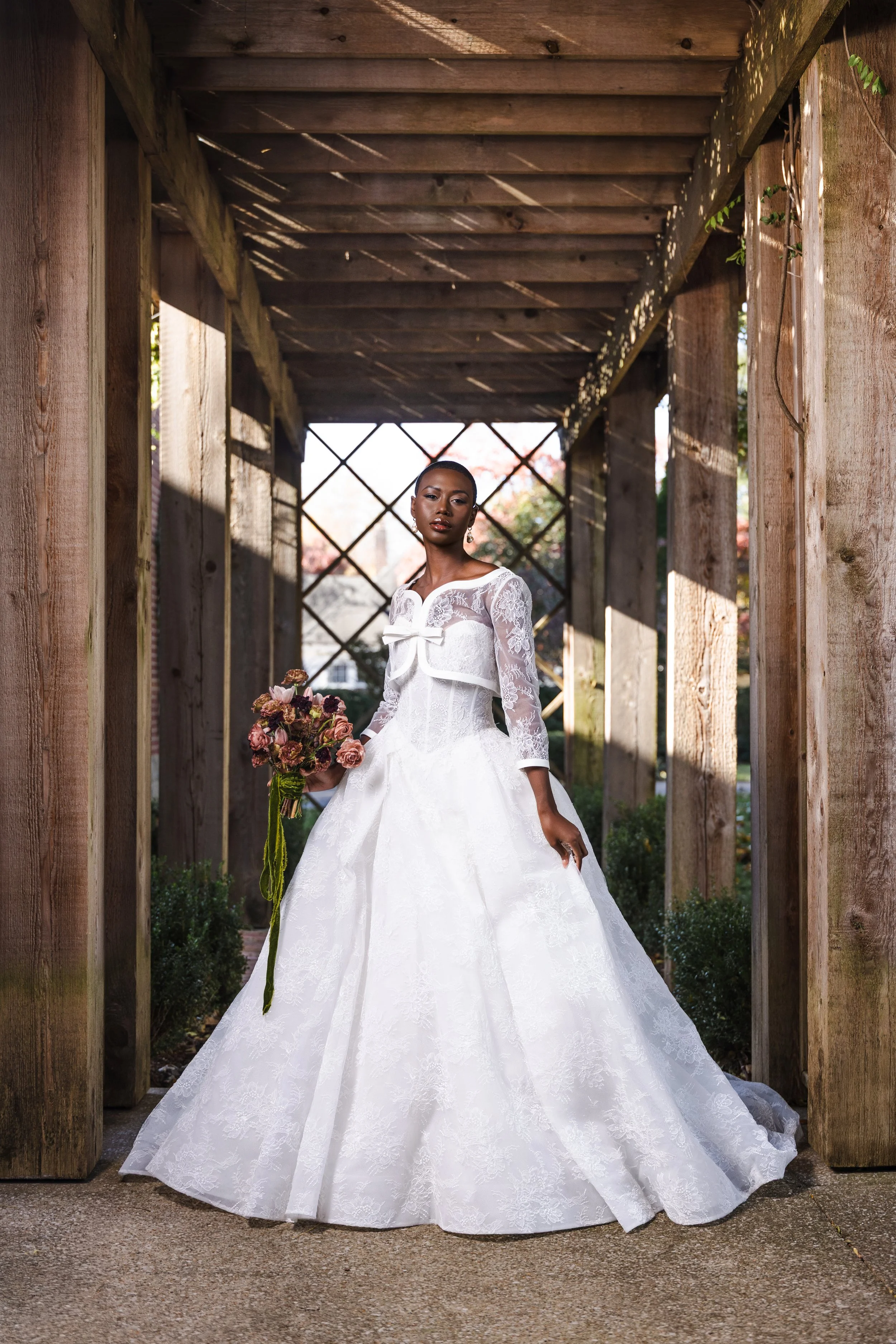 Full-length bridal portrait under rustic trellis with diagonal sunlight, showcasing romantic lace gown and earthy-toned bouquet