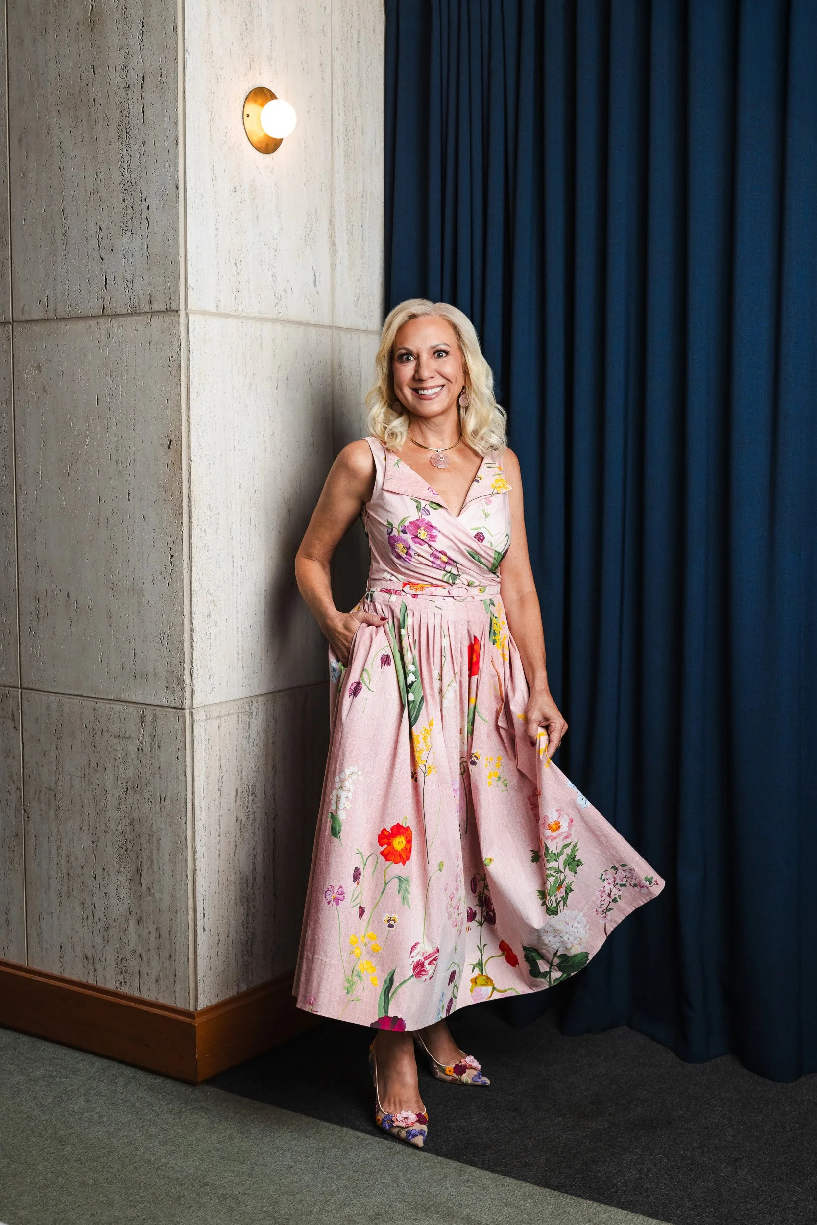 Julia Baker in a blush floral wrap dress, leaning against a travertine stone wall for her Nashville editorial portrait.