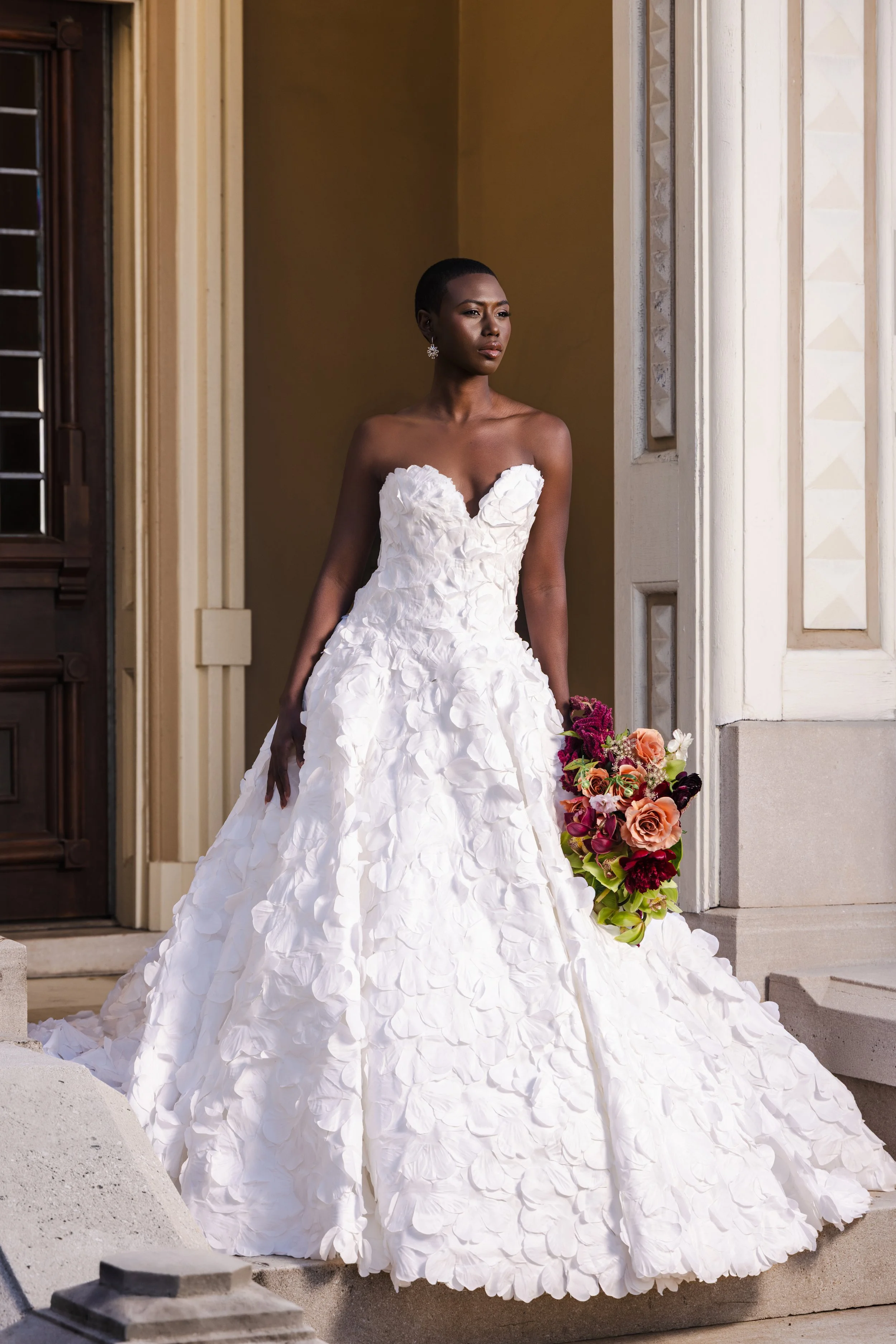 Bride in a strapless lace ball gown holding a colorful bouquet, standing in a grand doorway with classic architectural details for an nFocus wedding editorial.