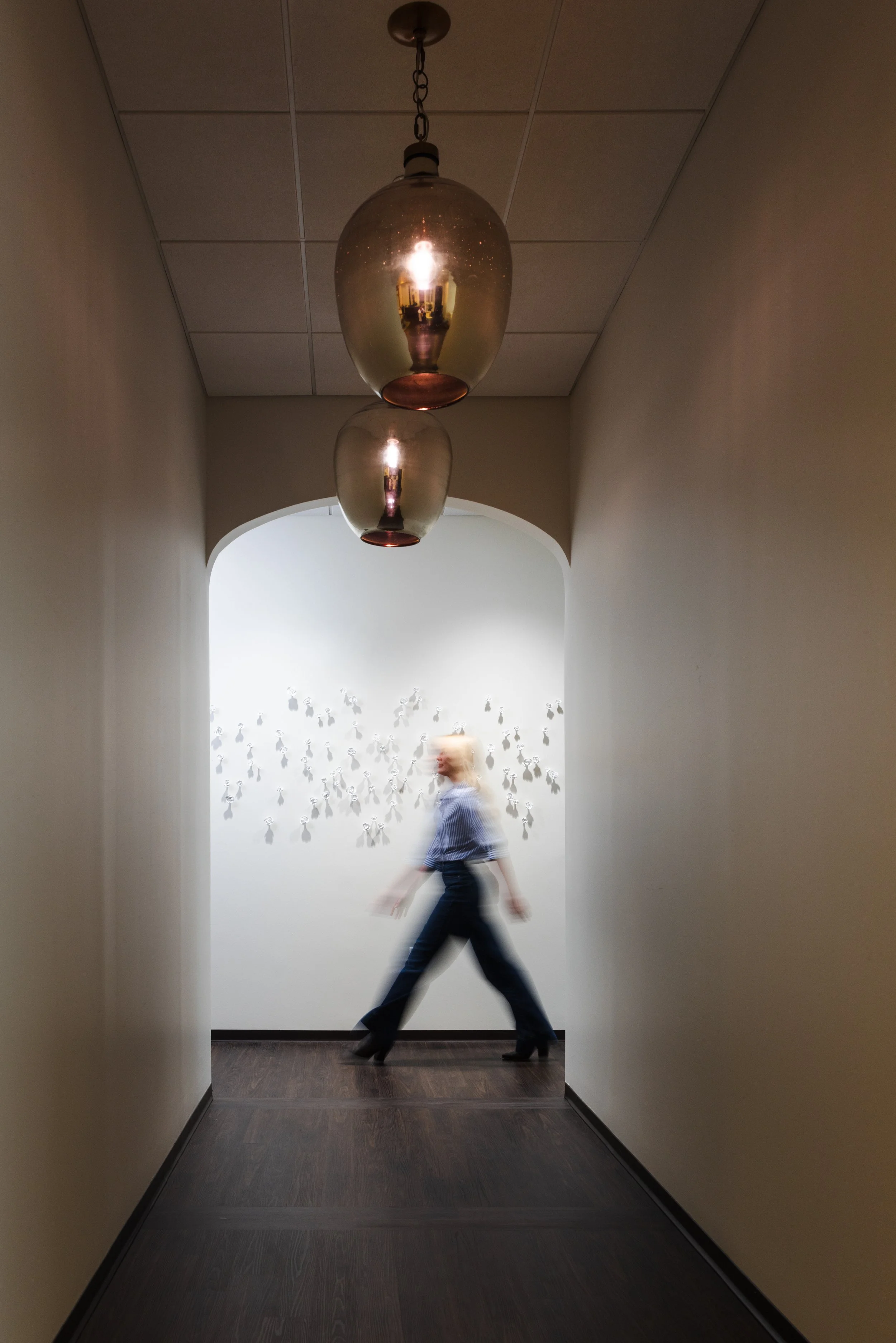 Blurred motion shot of a woman walking through an arched hallway beneath glass pendant lights in a cosmetic surgery office.