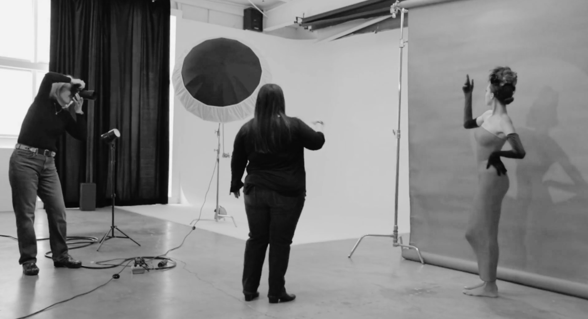 Behind the scenes black and white photo of a photographer directing a studio portrait session with lighting equipment and model.
