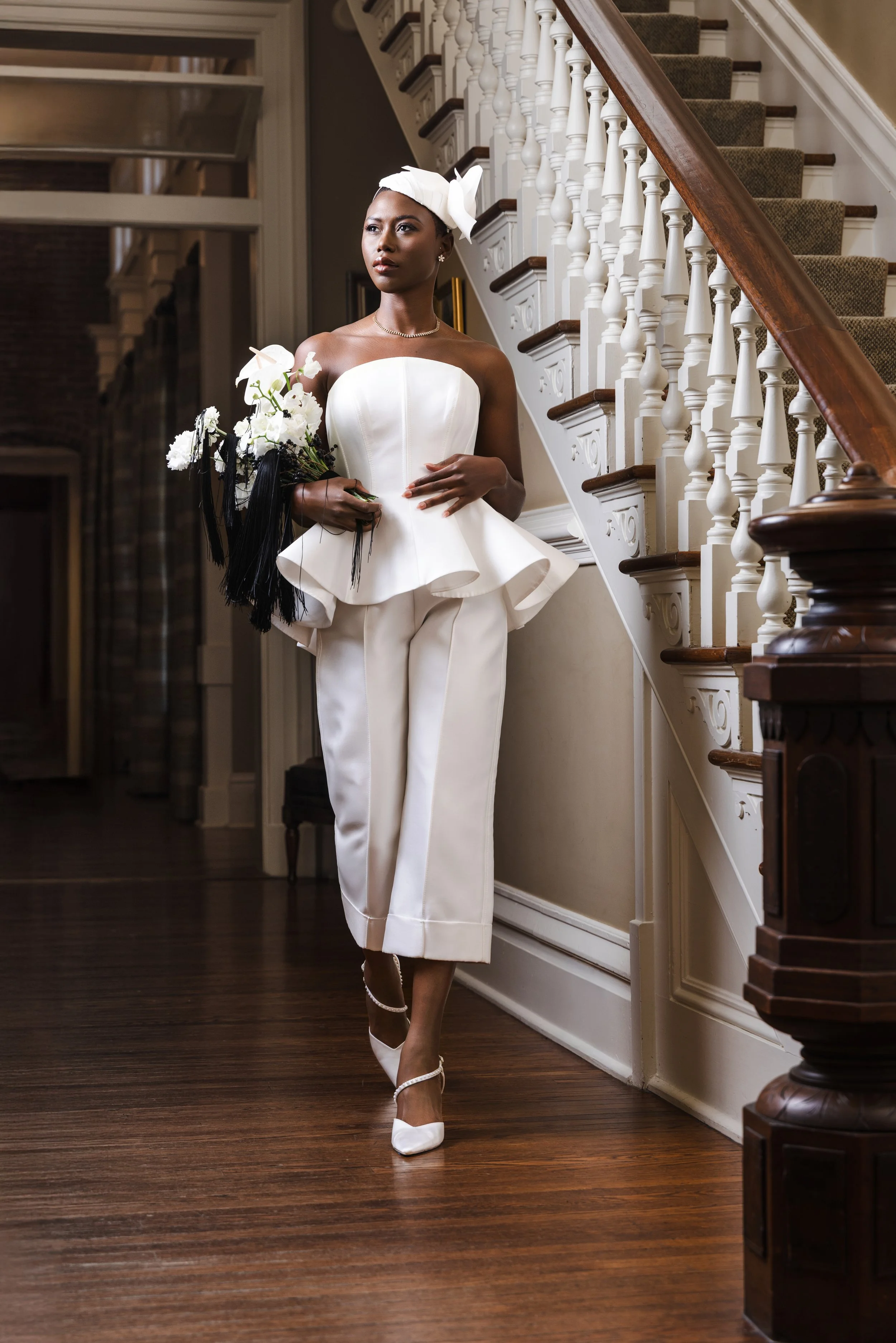 Bride wearing a modern strapless white bridal pantsuit with a sculptural peplum top and cropped trousers, styled with a minimalist headpiece and pointed heels, holding a bouquet with black ribbon details while walking through an elegant historic inte