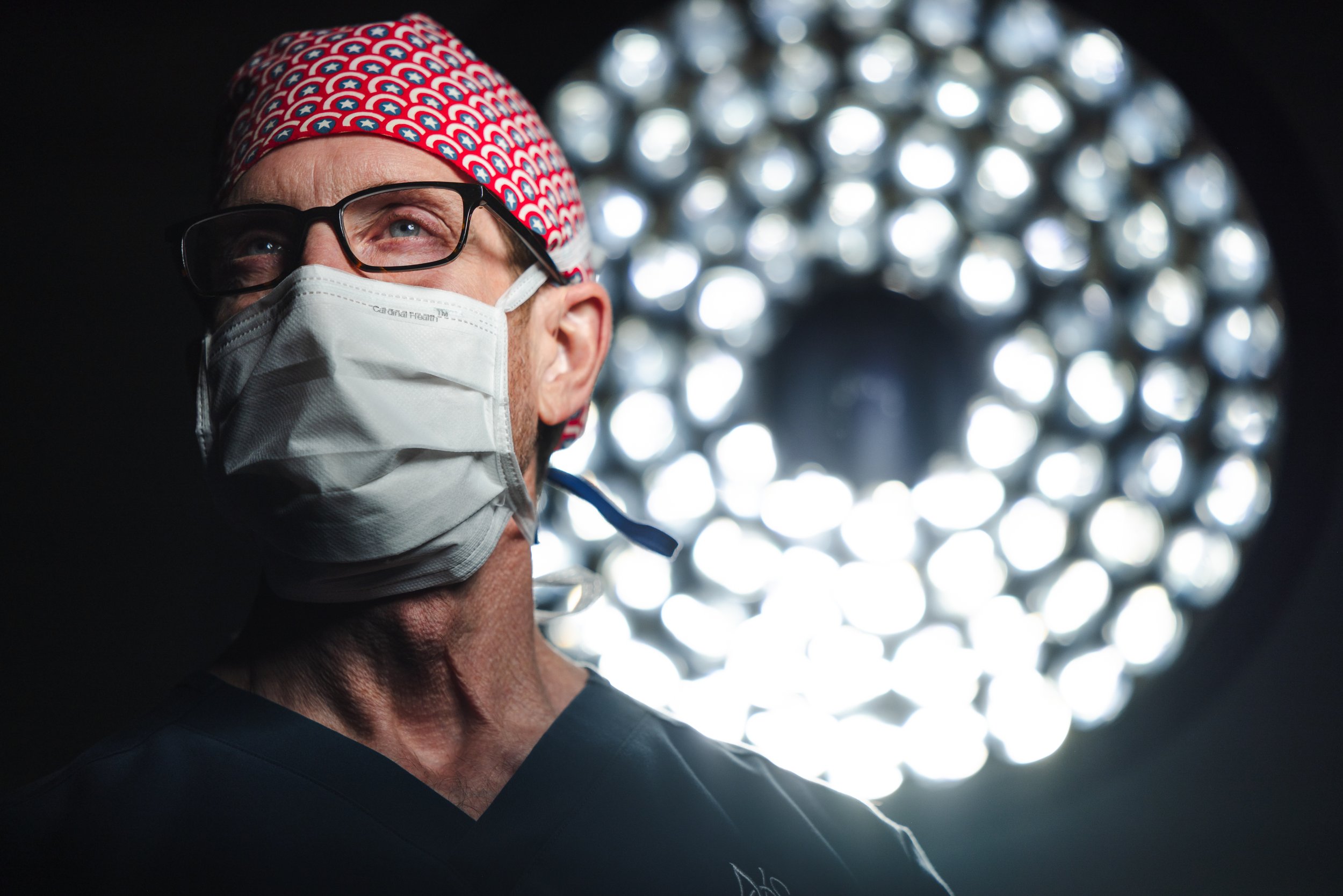 Surgeon in a red patterned scrub cap and surgical mask standing beneath a round operating room light.