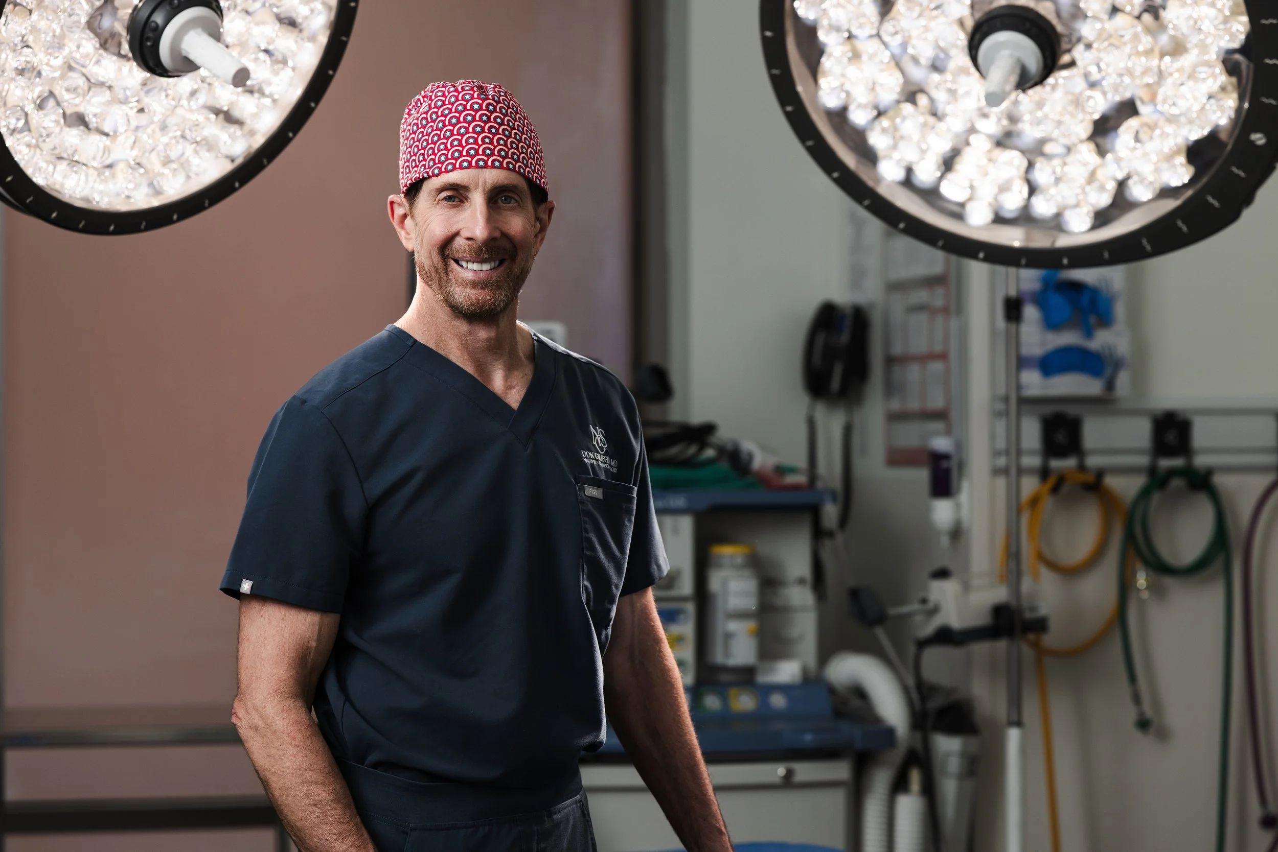 Surgeon in navy scrubs and a red patterned cap smiling under operating room lights, his practice name embroidered on his scrubs.