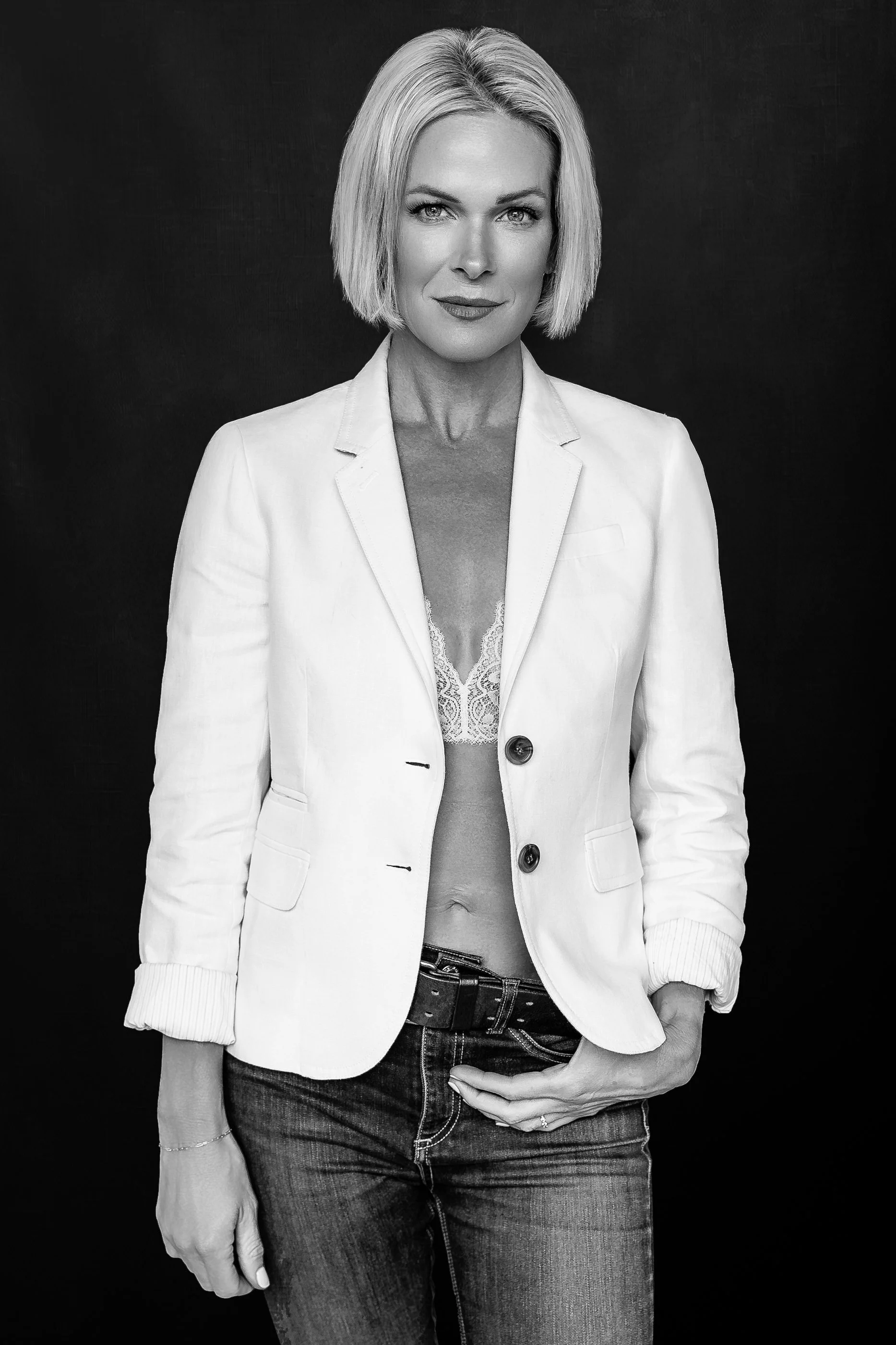 A striking black and white creative portrait of a confident blonde woman standing against a dark studio backdrop. Styled in a white blazer layered over delicate lace detailing with relaxed denim, the portrait blends modern fashion-inspired styling wi