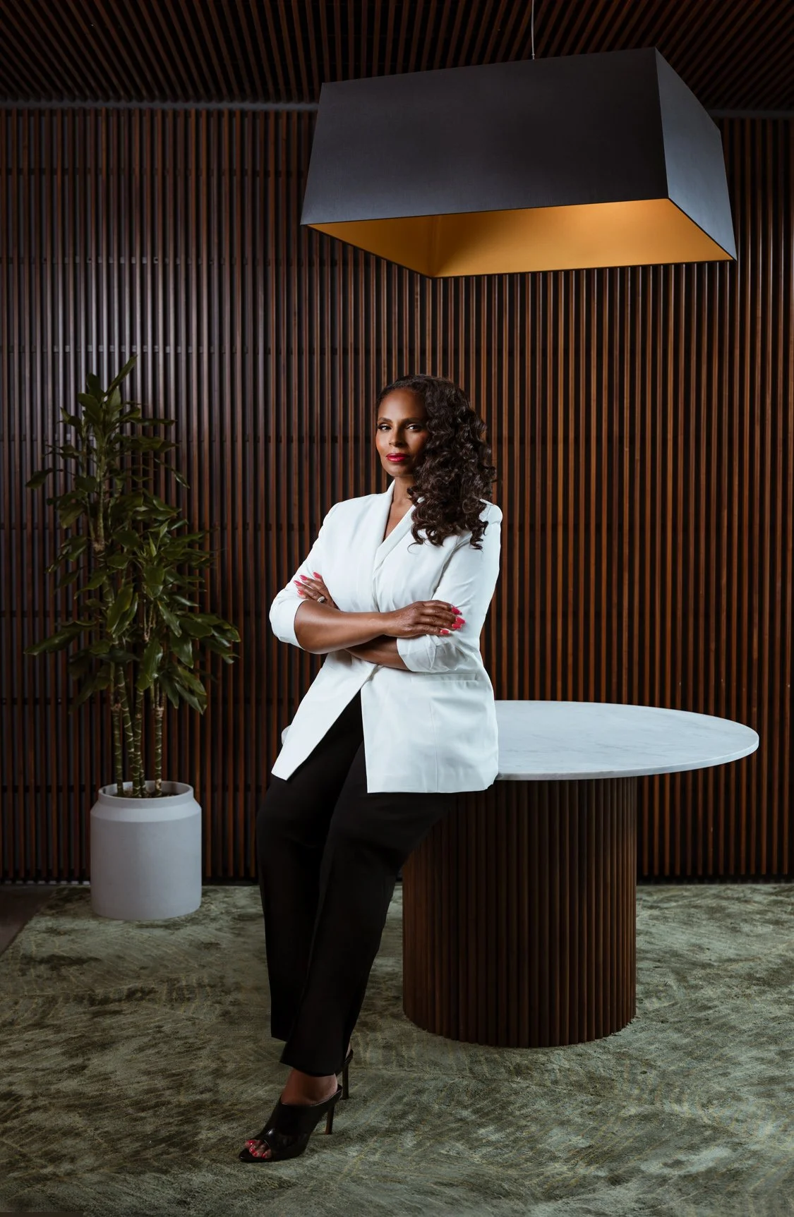 Executive branding portrait of a woman in a white blazer with arms crossed standing beside a marble table in a modern office with wood slat walls, photographed by Tausha Dickinson in Nashville