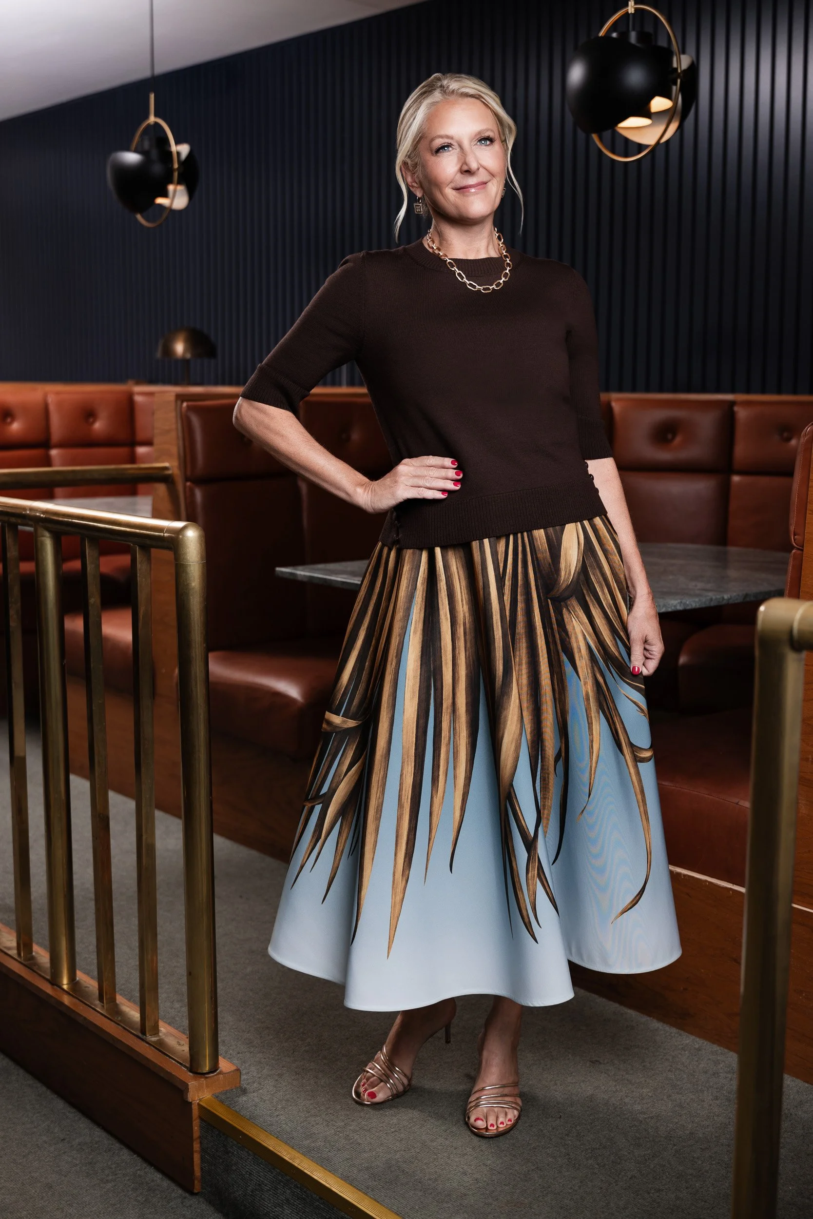 Caroline Cook wearing a brown sweater and palm print skirt, photographed at a brass bar railing in Nashville for nFocus Magazine's Model Behavior 2026.