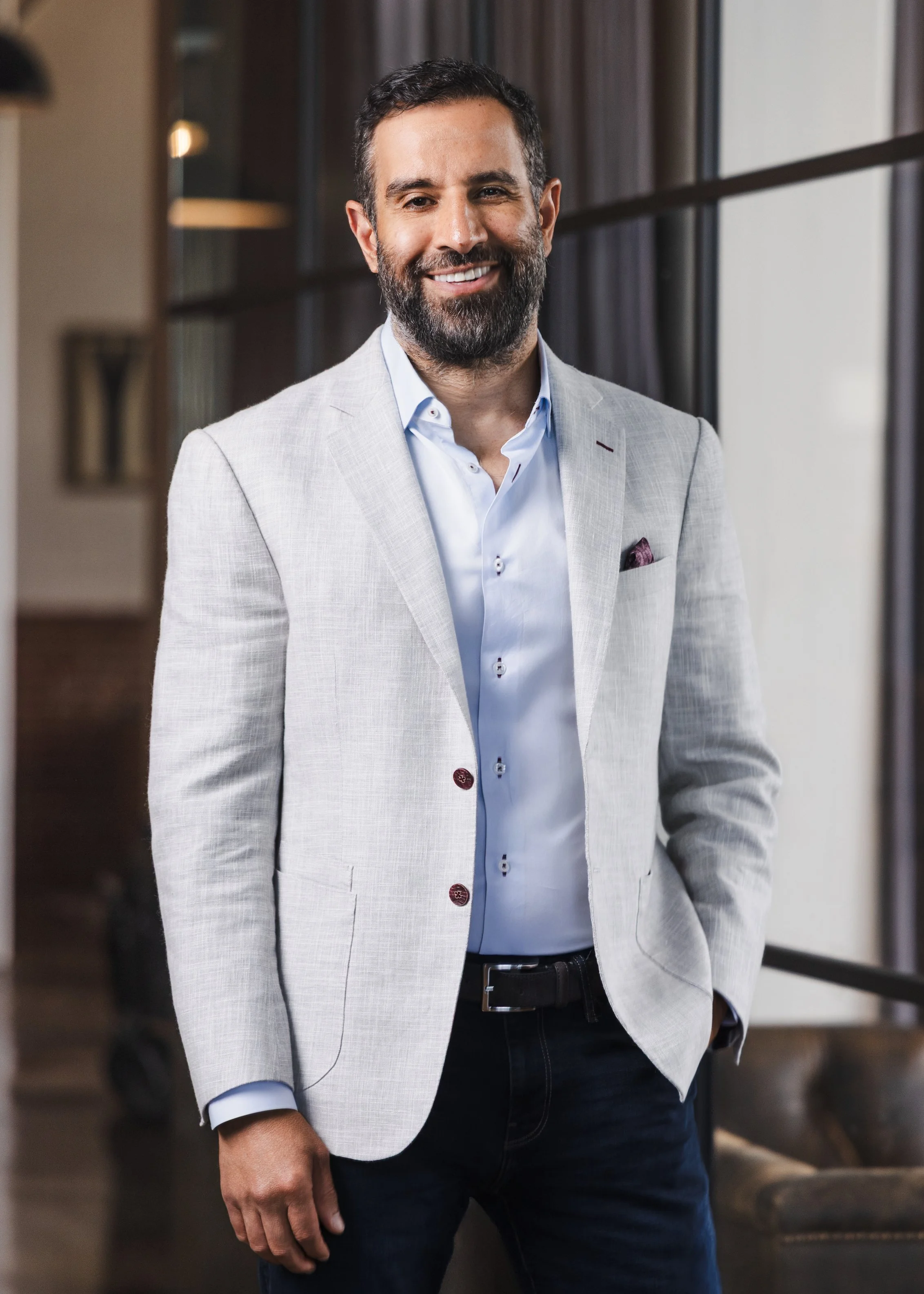 Professional headshot of a smiling bearded man in a light gray blazer standing in a warmly lit upscale interior, photographed by Tausha Dickinson in Nashville
