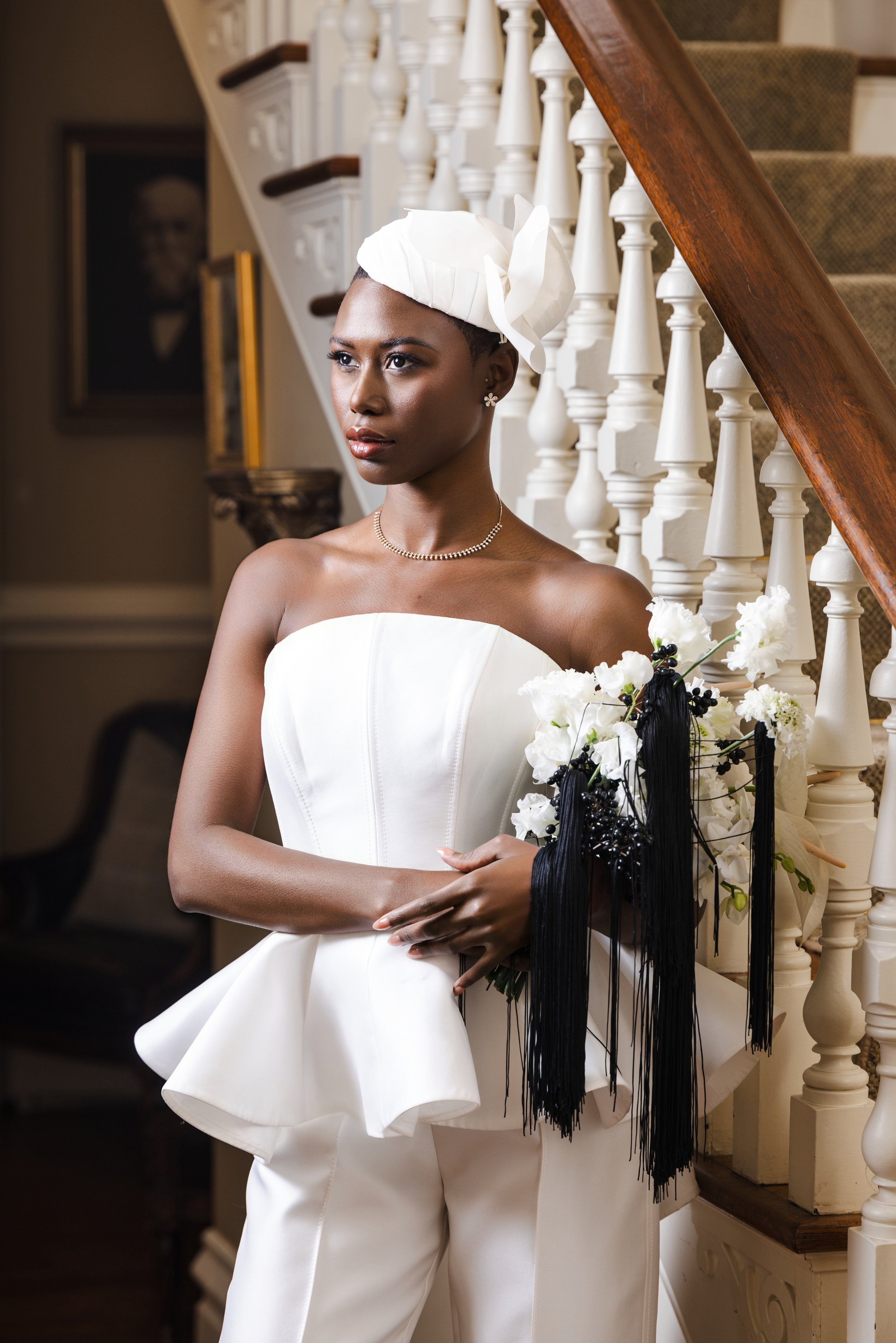 Bride in a modern strapless white jumpsuit with a sculptural peplum detail, holding a bouquet beside an ornate staircase, featured as the nFocus January 2026 cover image.