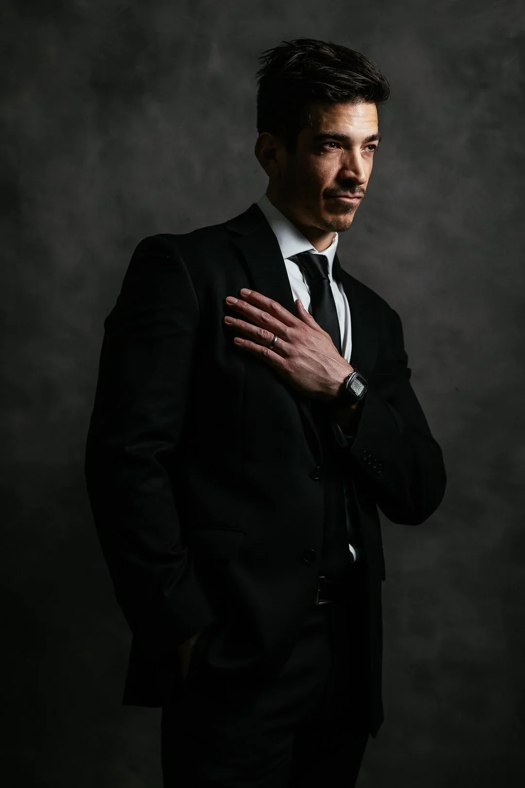 Professional event portrait of man in black suit and white shirt with hand on chest, dramatic studio lighting.