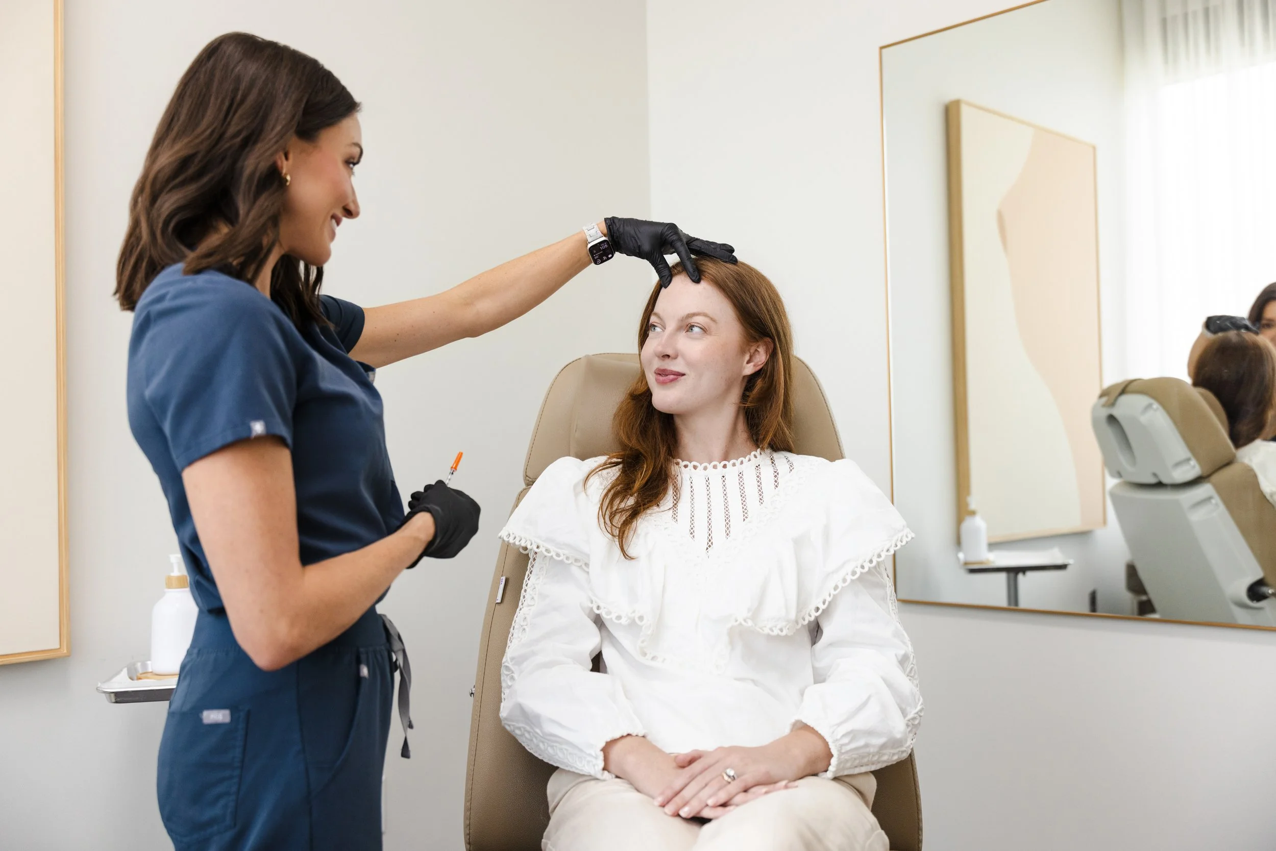 Injector in navy scrubs administering a forehead injection to a smiling redheaded patient in a treatment room.
