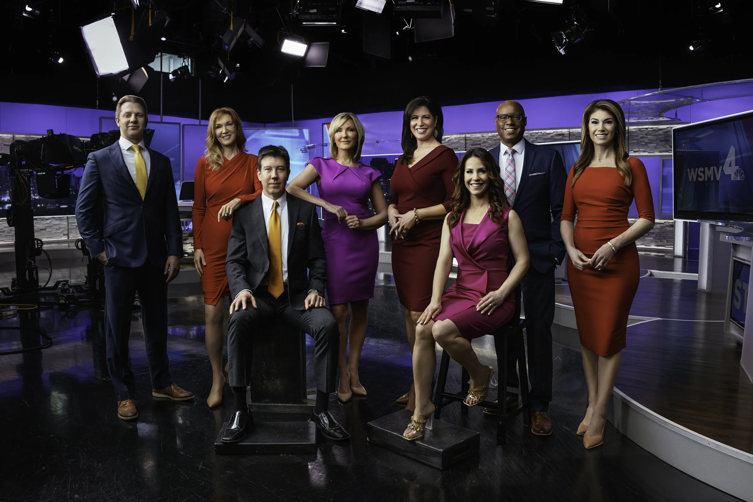 Group of nine news anchors and journalists in a television news studio.