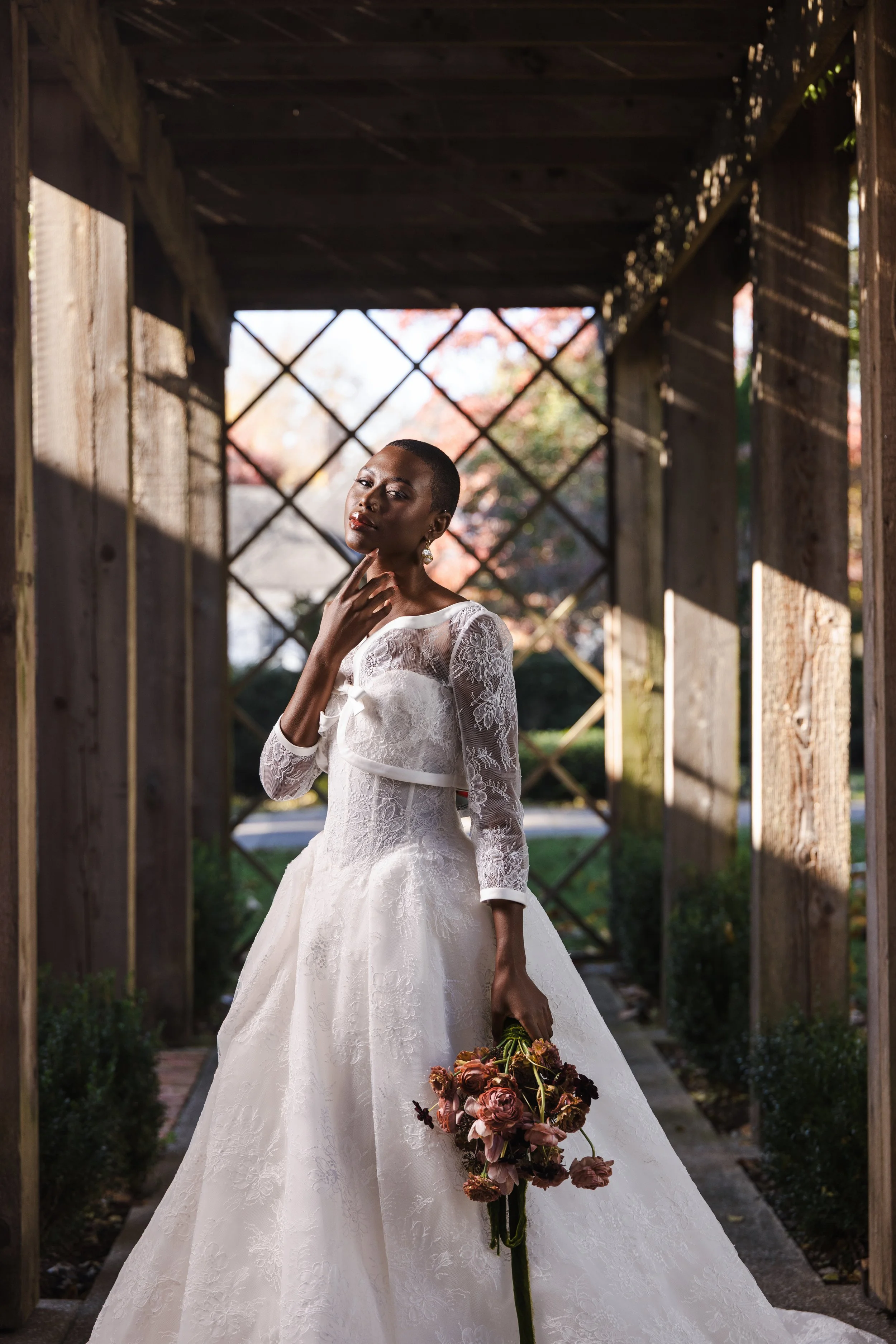 Bride in lace ballgown poses beneath wooden trellis in morning light, holding a bouquet of muted florals with soft shadows across her face