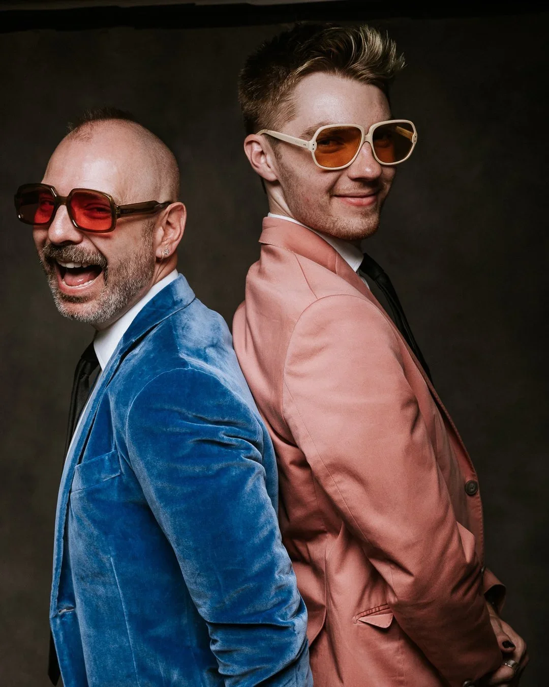 Murder mystery event portrait of two men in colorful sunglasses and velvet blazers posing back to back with playful expressions.