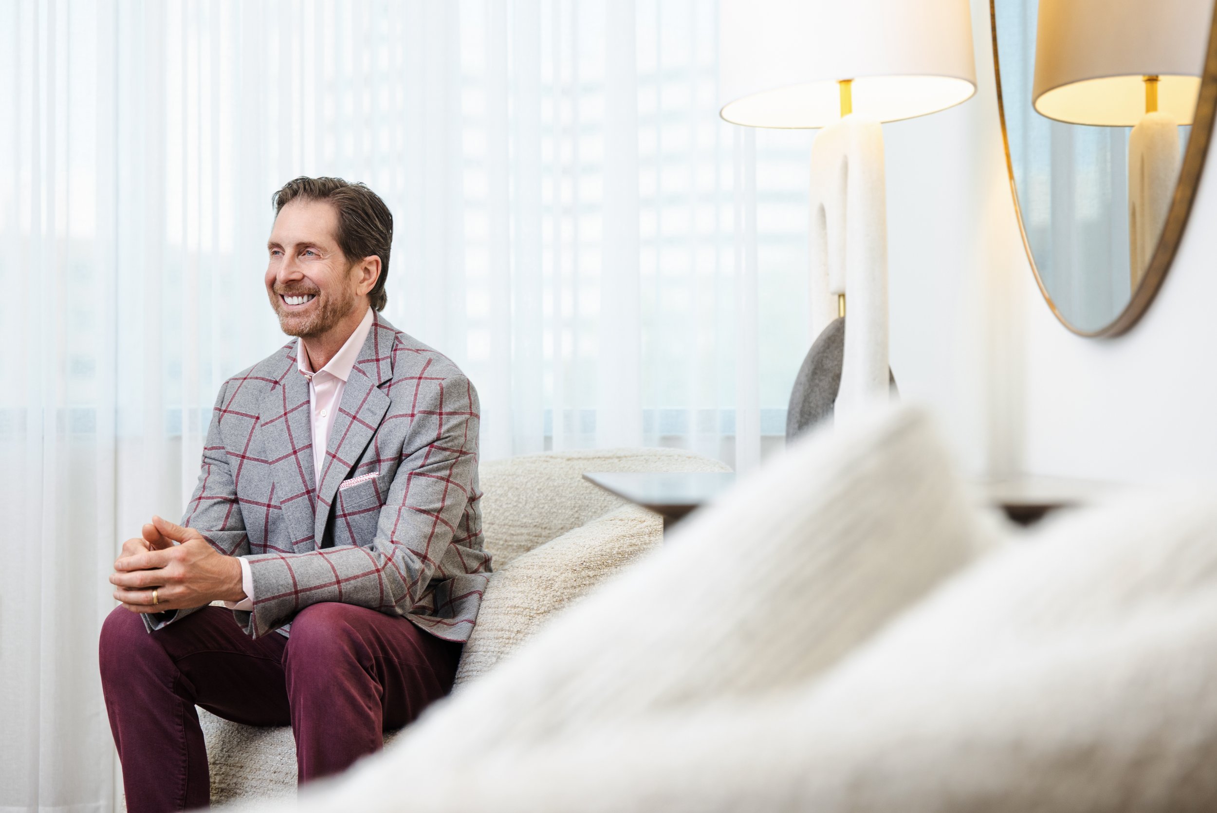 Man in a gray and red plaid blazer and burgundy pants smiling while seated on a couch in a stylish lounge area.