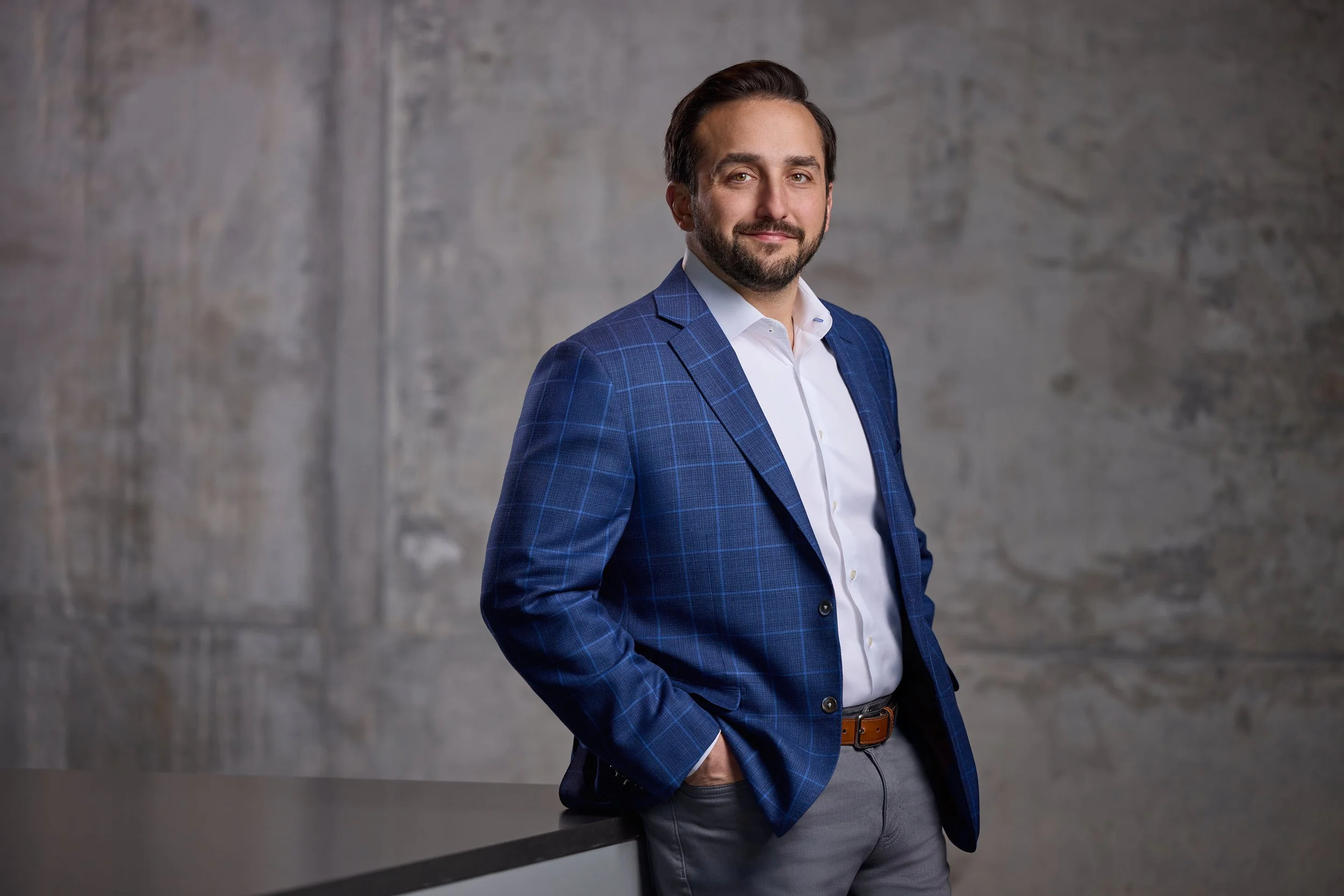 Professional headshot of a bearded man in a blue plaid blazer leaning against a concrete textured wall, photographed by Tausha Dickinson Photography in Nashville