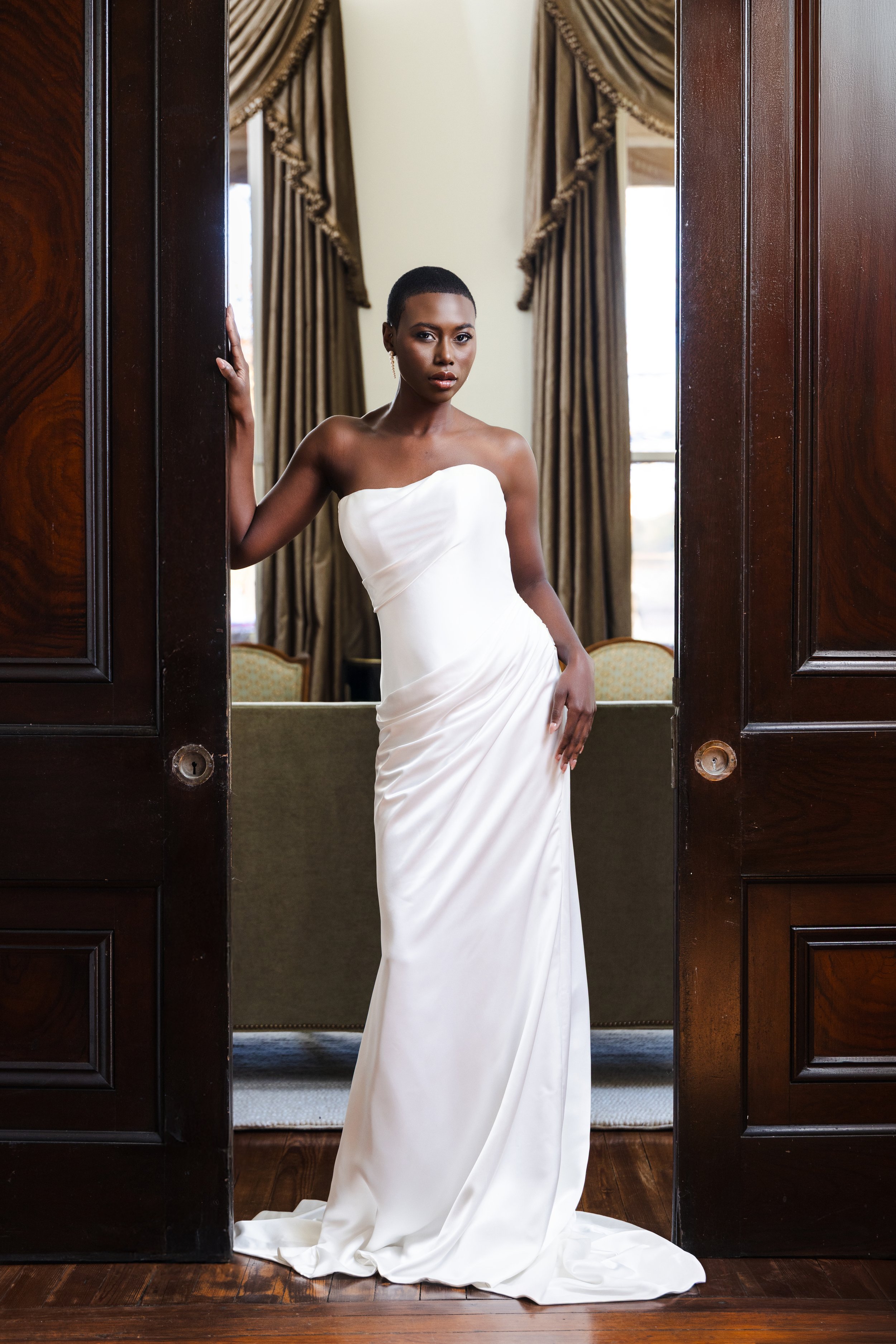 Bride in a structured sculptural gown stands confidently in front of ornate wood doors and dramatic drapery, lit with soft bounced light for added depth