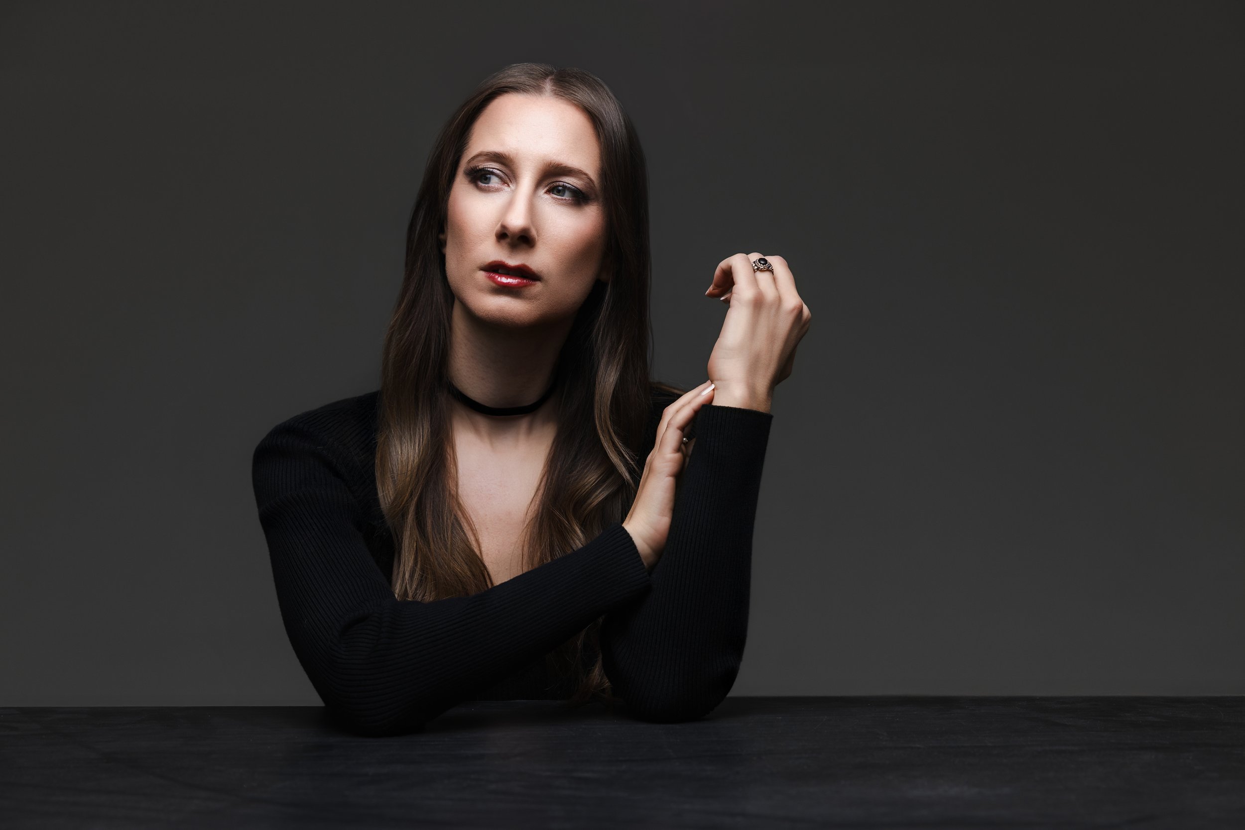 Moody studio portrait of Nashville singer-songwriter Lauren River resting her arms on a table, lit with soft overhead umbrella lighting against a dark gray background