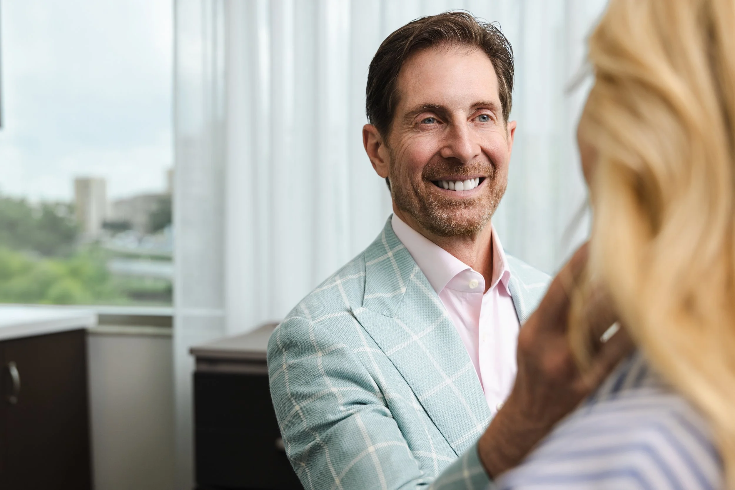 Close-up of a smiling man in a mint windowpane blazer during a consultation with a patient in the foreground.