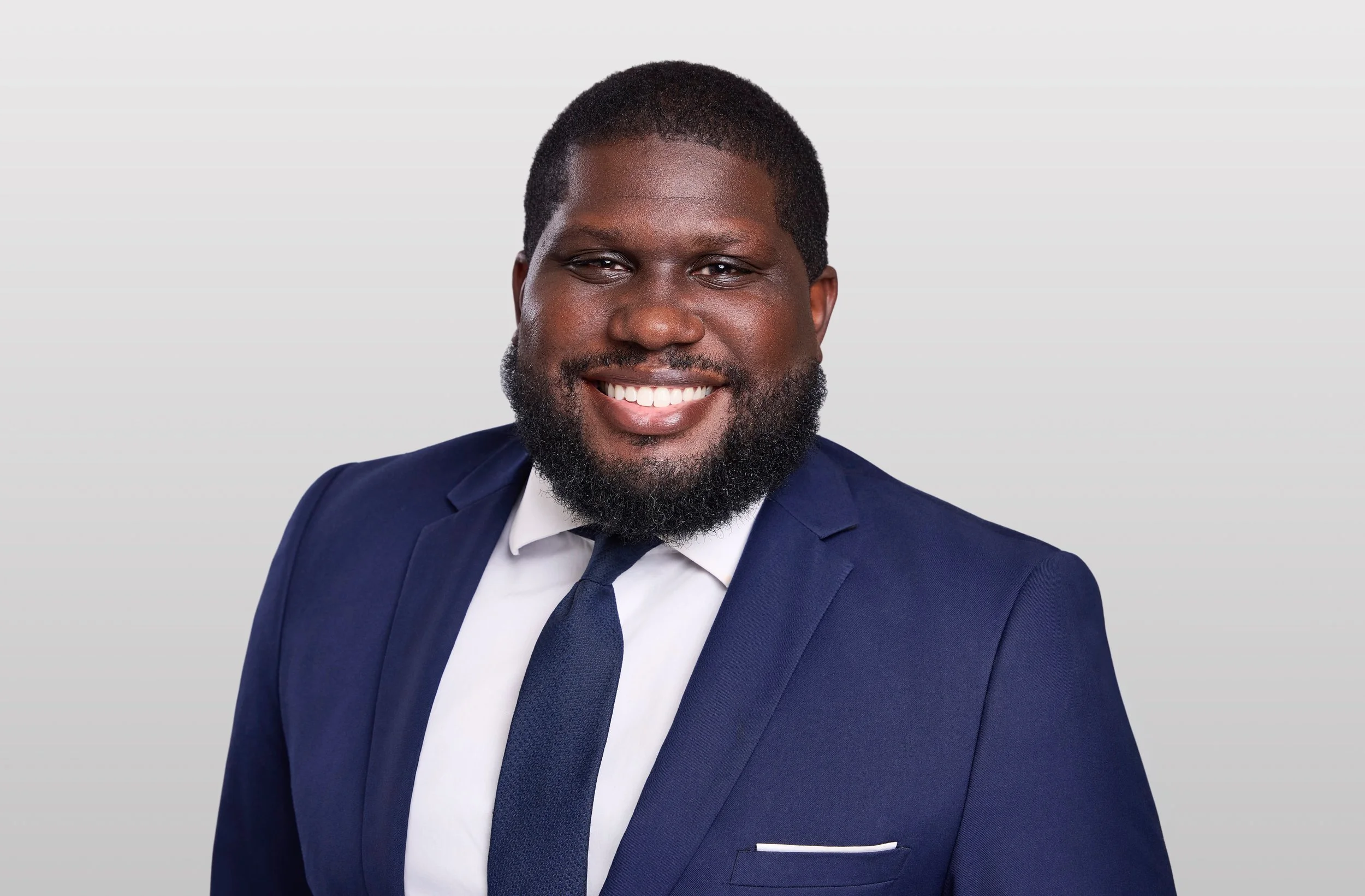 Business headshot of a smiling man in a navy suit and tie against a light gray background.