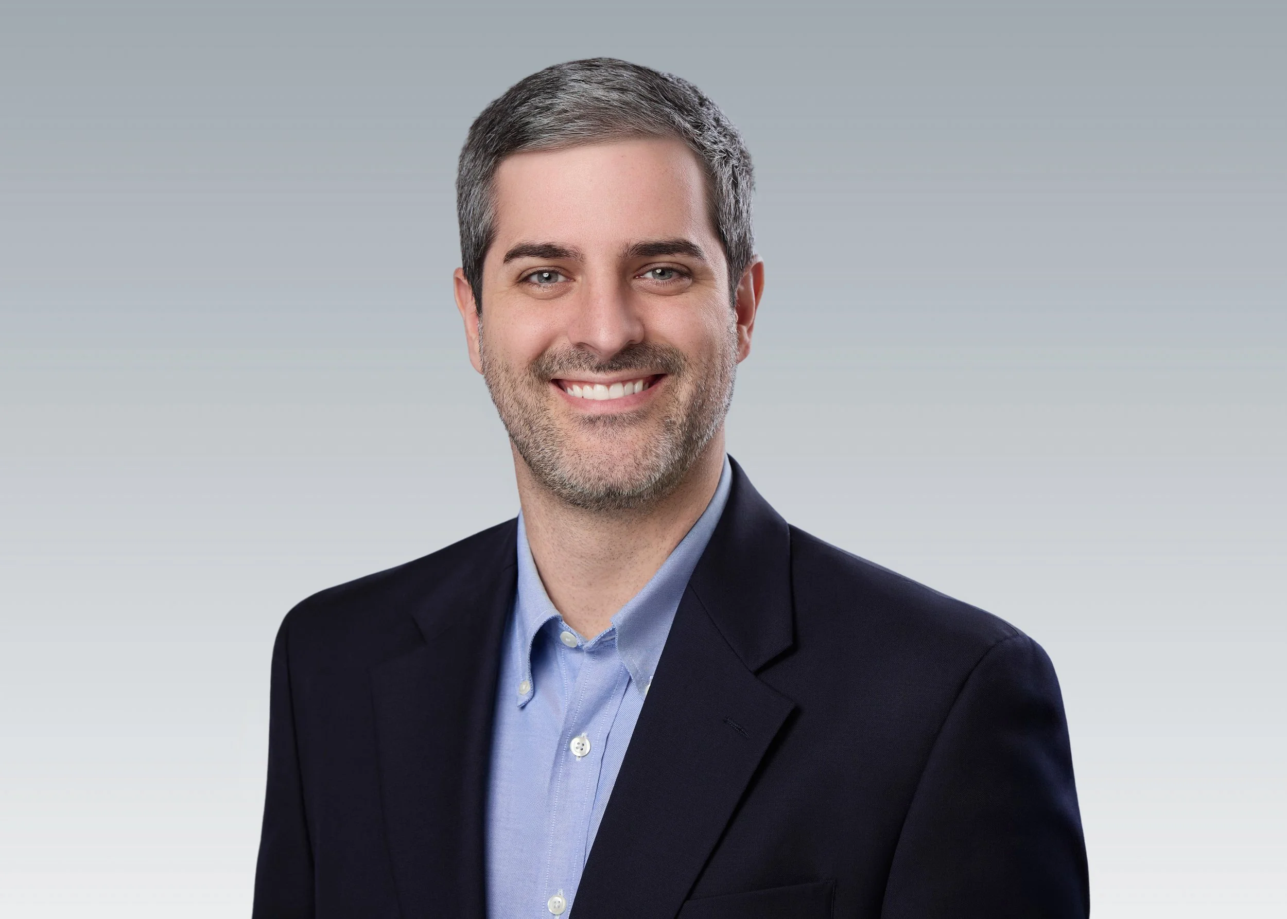 Executive team headshot of a smiling man in a dark blazer and light blue shirt on a matching gray backdrop, part of a cohesive company headshot session by Tausha Dickinson, Franklin TN