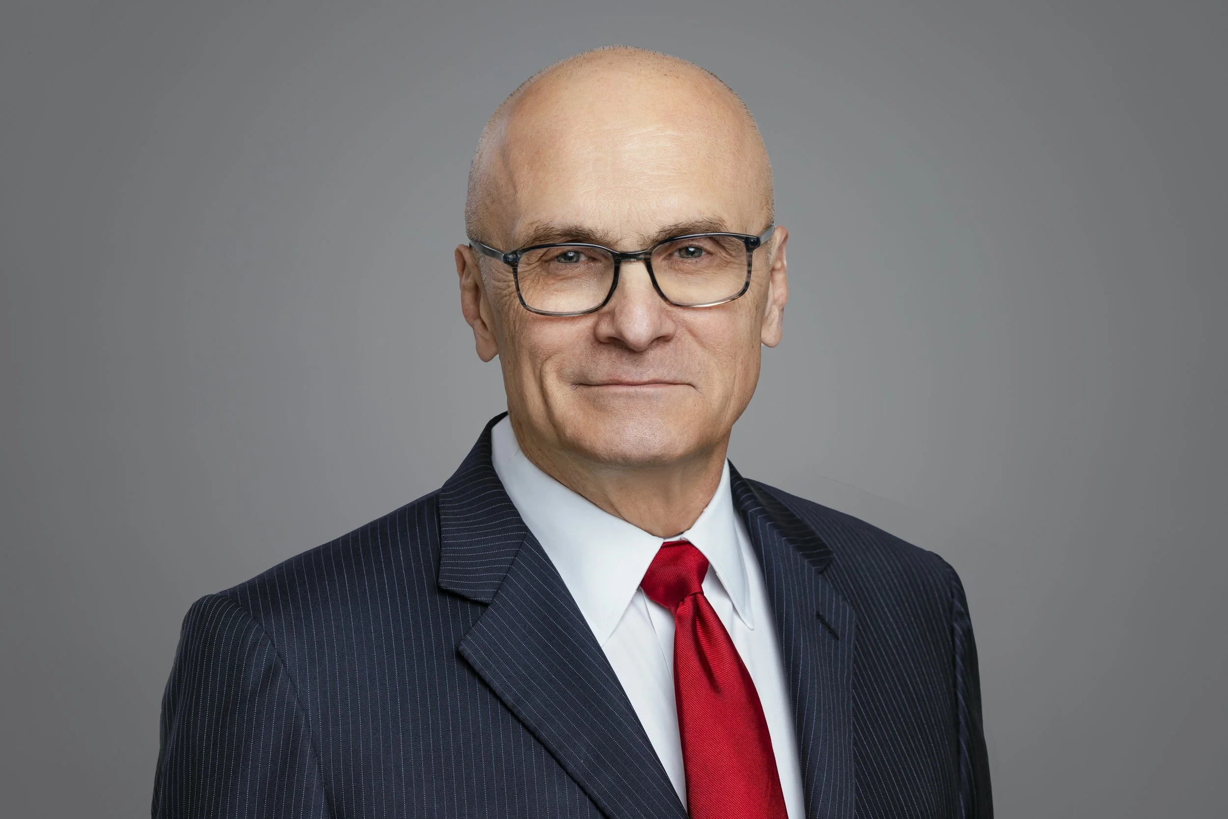 Executive headshot of a distinguished man with glasses in a dark suit and red tie against a gray background.