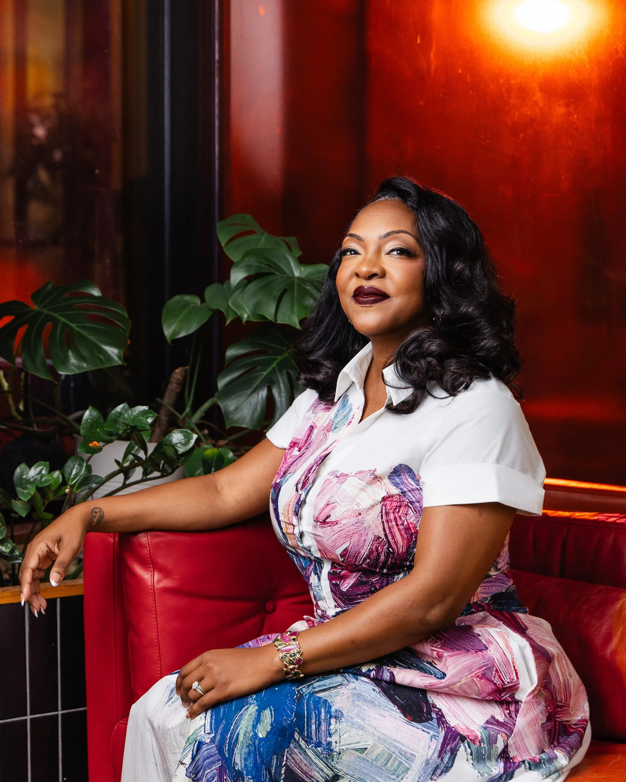 Christiane Buggs seated on a red leather couch in a painterly floral shirtdress for her Nashville editorial portrait session.
