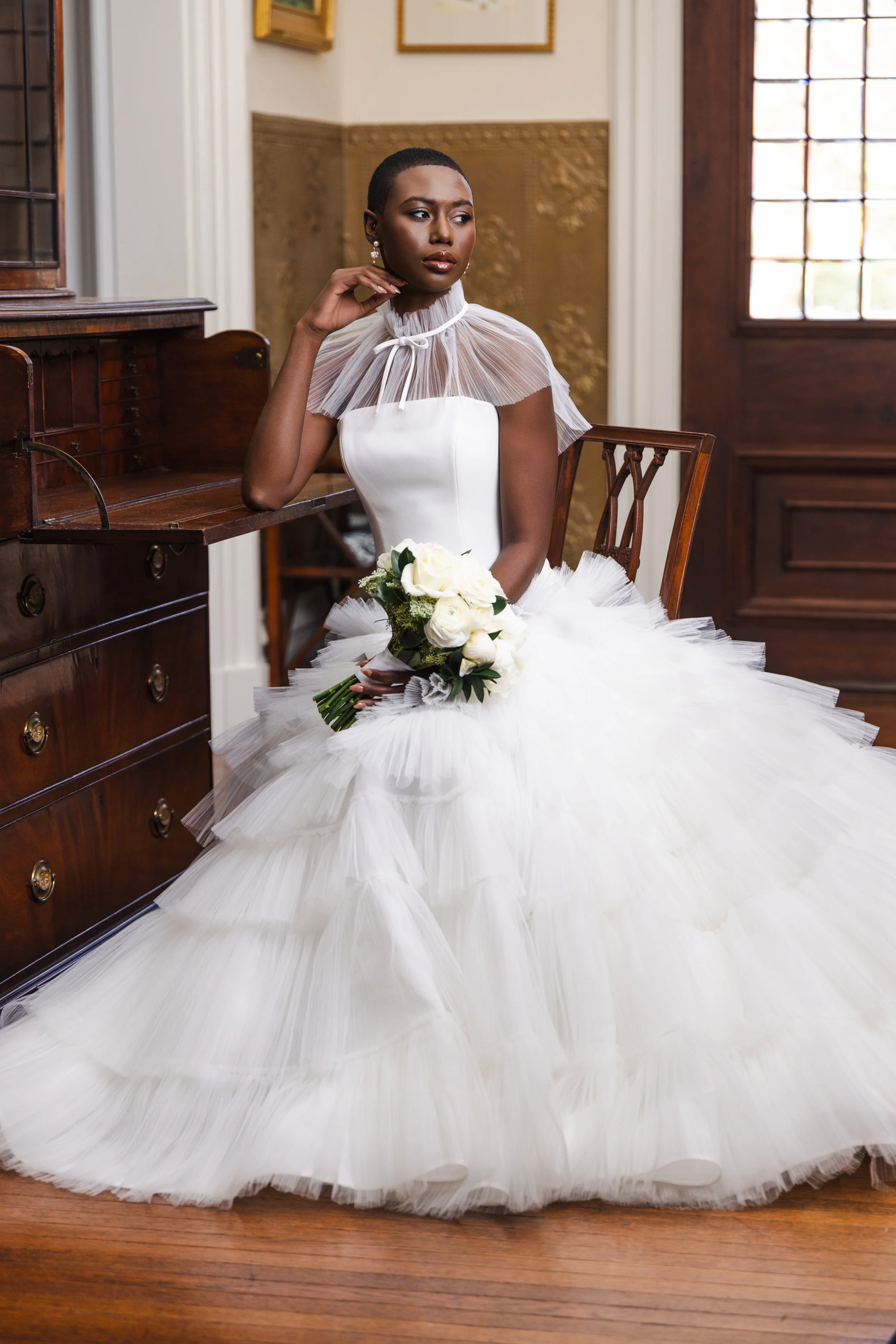 Bride in layered tulle gown and sheer capelet poses against textured wallpaper, styled for a modern yet romantic bridal editorial look