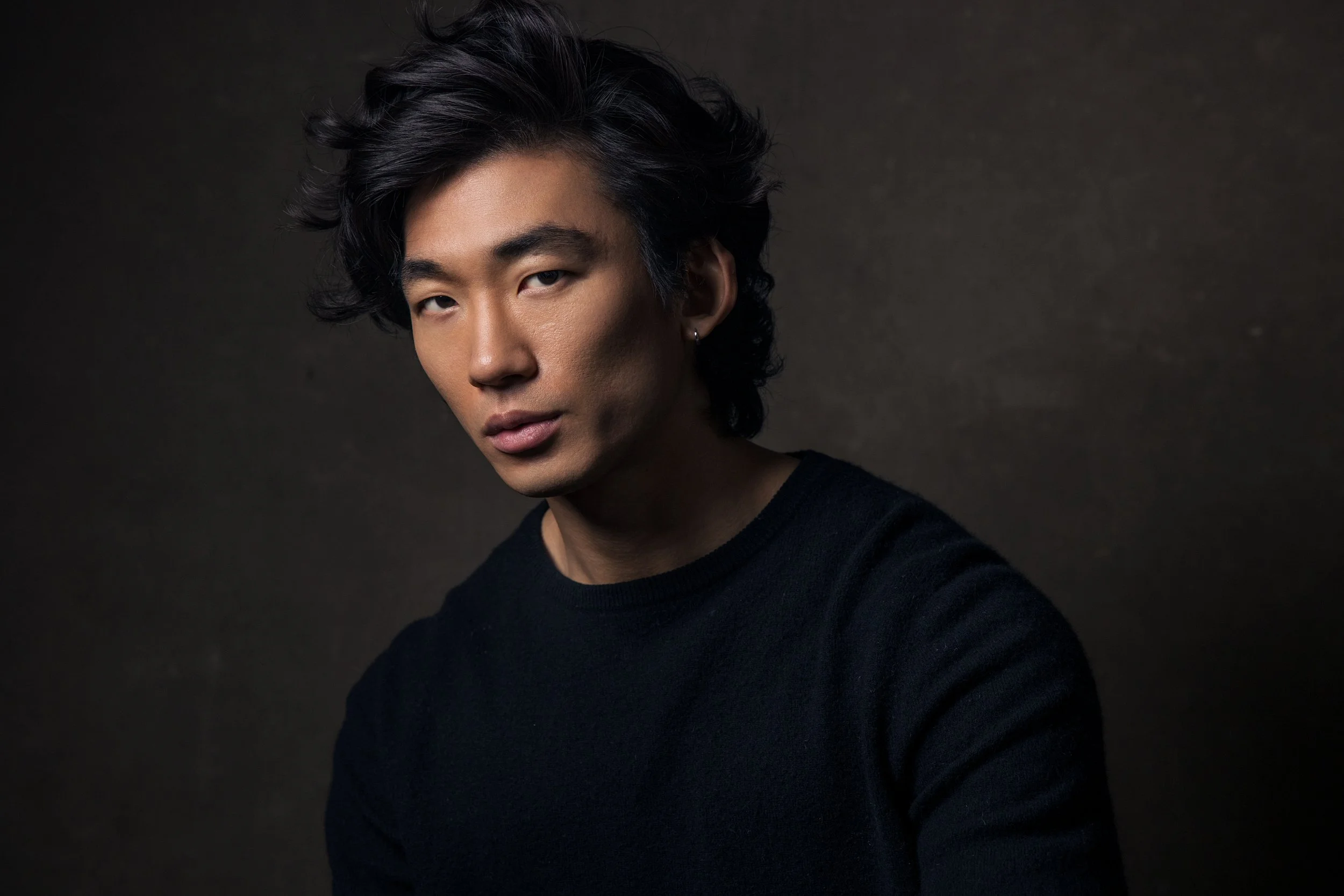 Studio headshot of young Asian man in black sweater against dark textured background, dramatic directional lighting, Nashville headshot photographer Tausha Dickinson